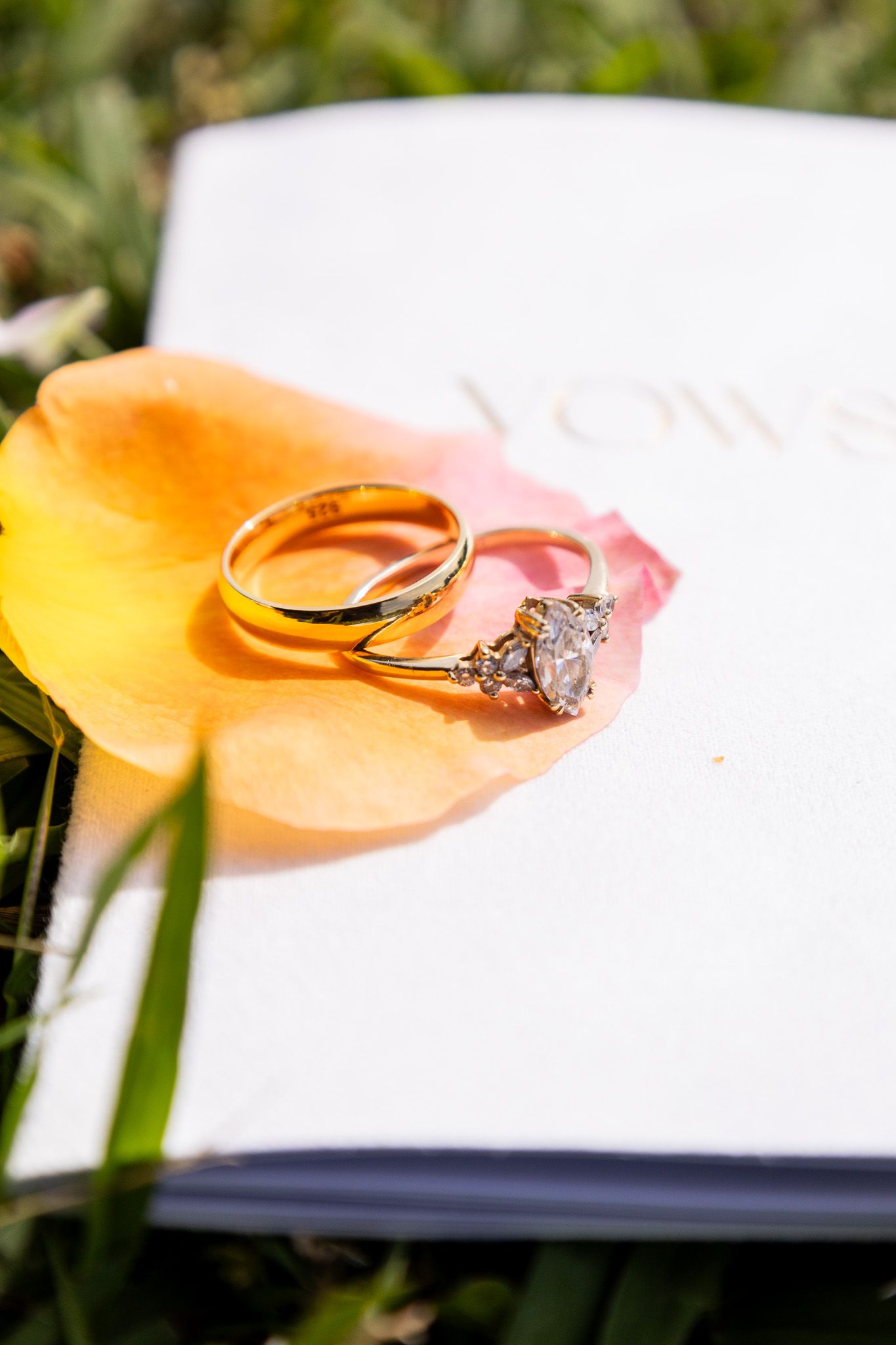 Two gold wedding rings resting on a yellow flower petal atop a white vows book outdoors.