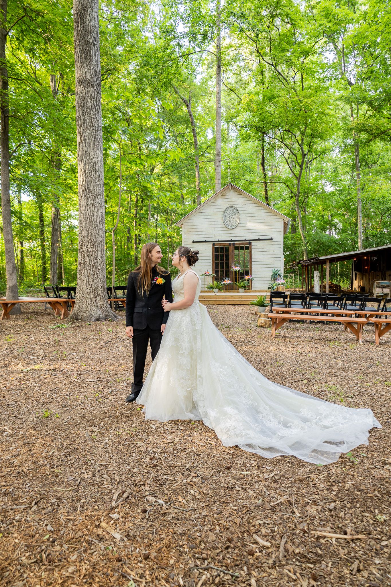 Bride and groom share a romantic moment in a wooded outdoor wedding venue with a white chapel in the background.