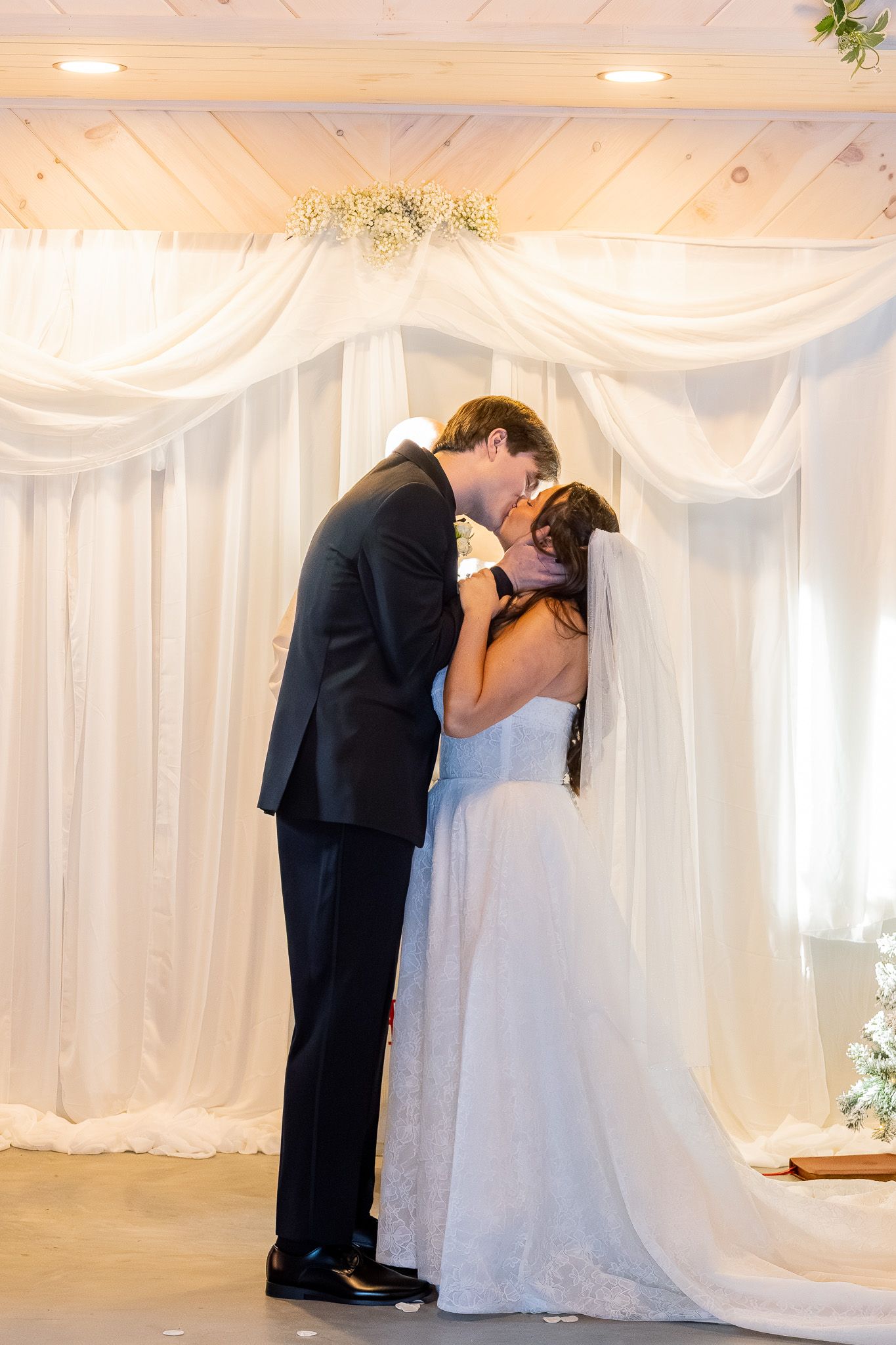 Bride and groom share first kiss at altar adorned with white draping and baby's breath floral decor.