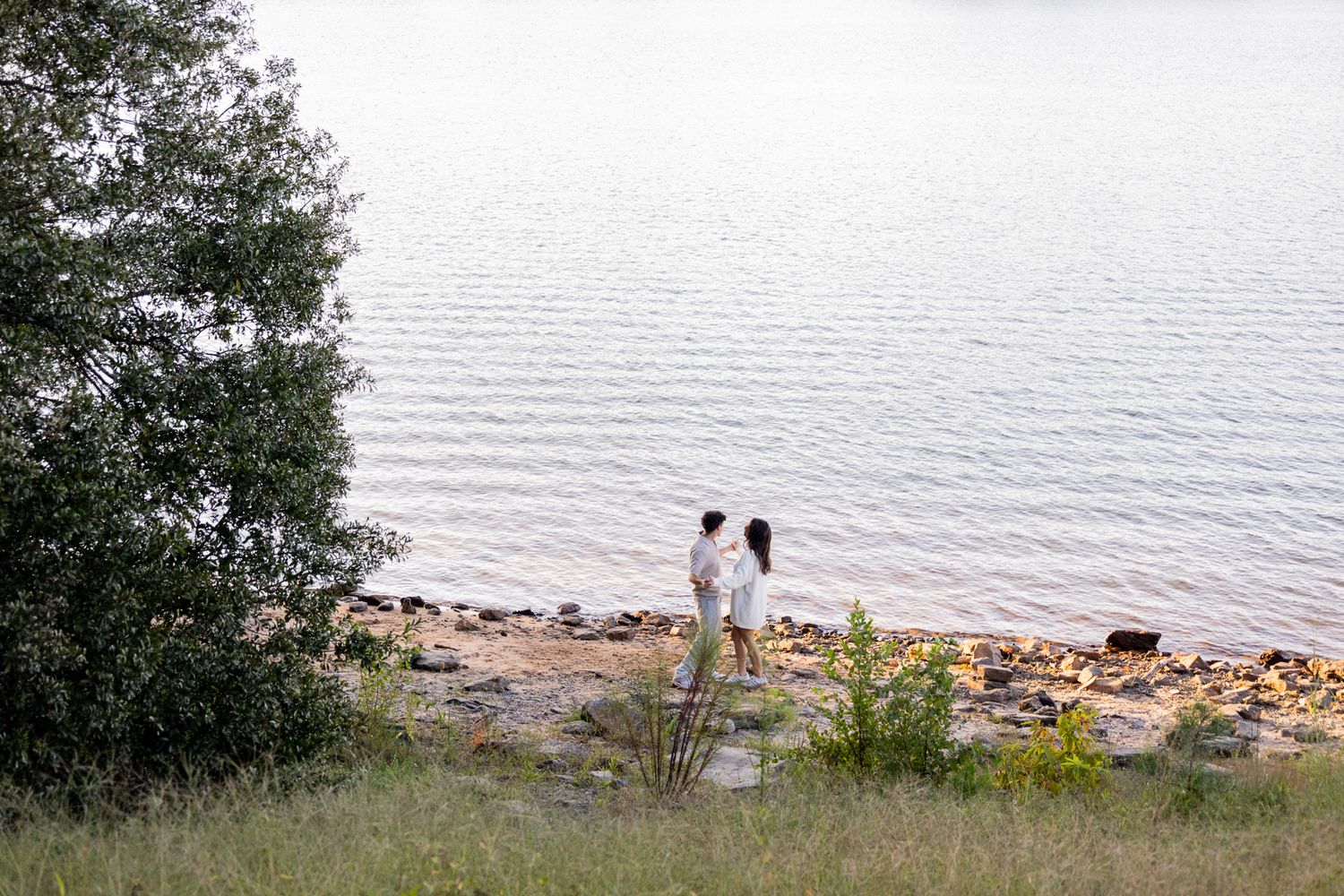 Couple standing together on a rocky shoreline by a calm lake, surrounded by lush greenery and trees.