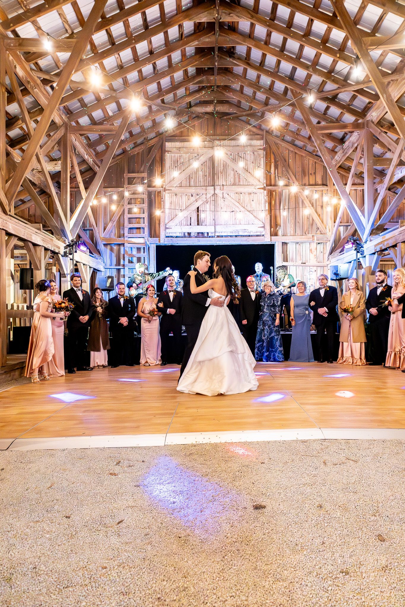Bride and groom share first dance in a rustic barn venue with string lights and guests watching.