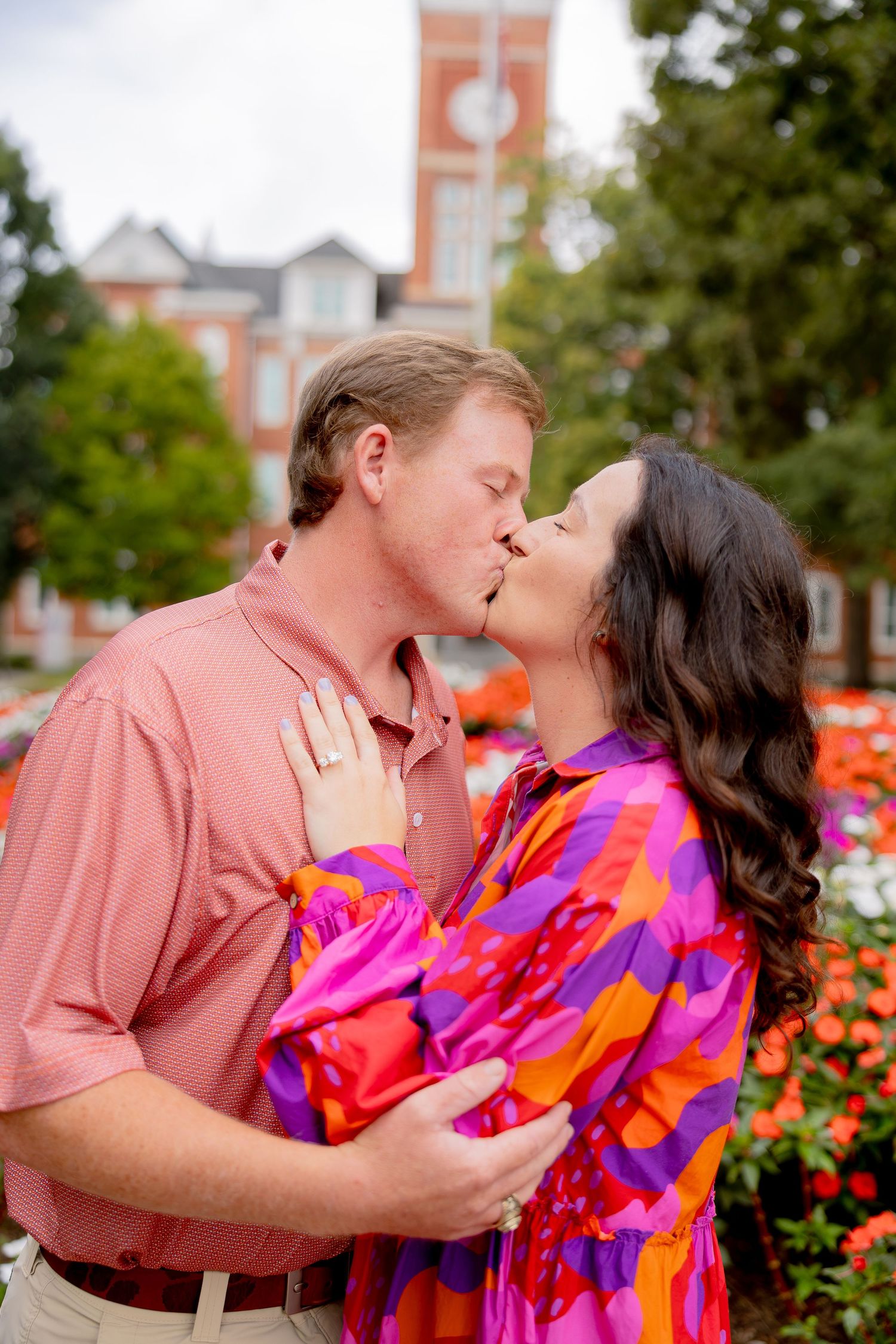 Couple sharing a romantic kiss outdoors near colorful flowers, woman wearing vibrant pink dress, man in coral shirt.
