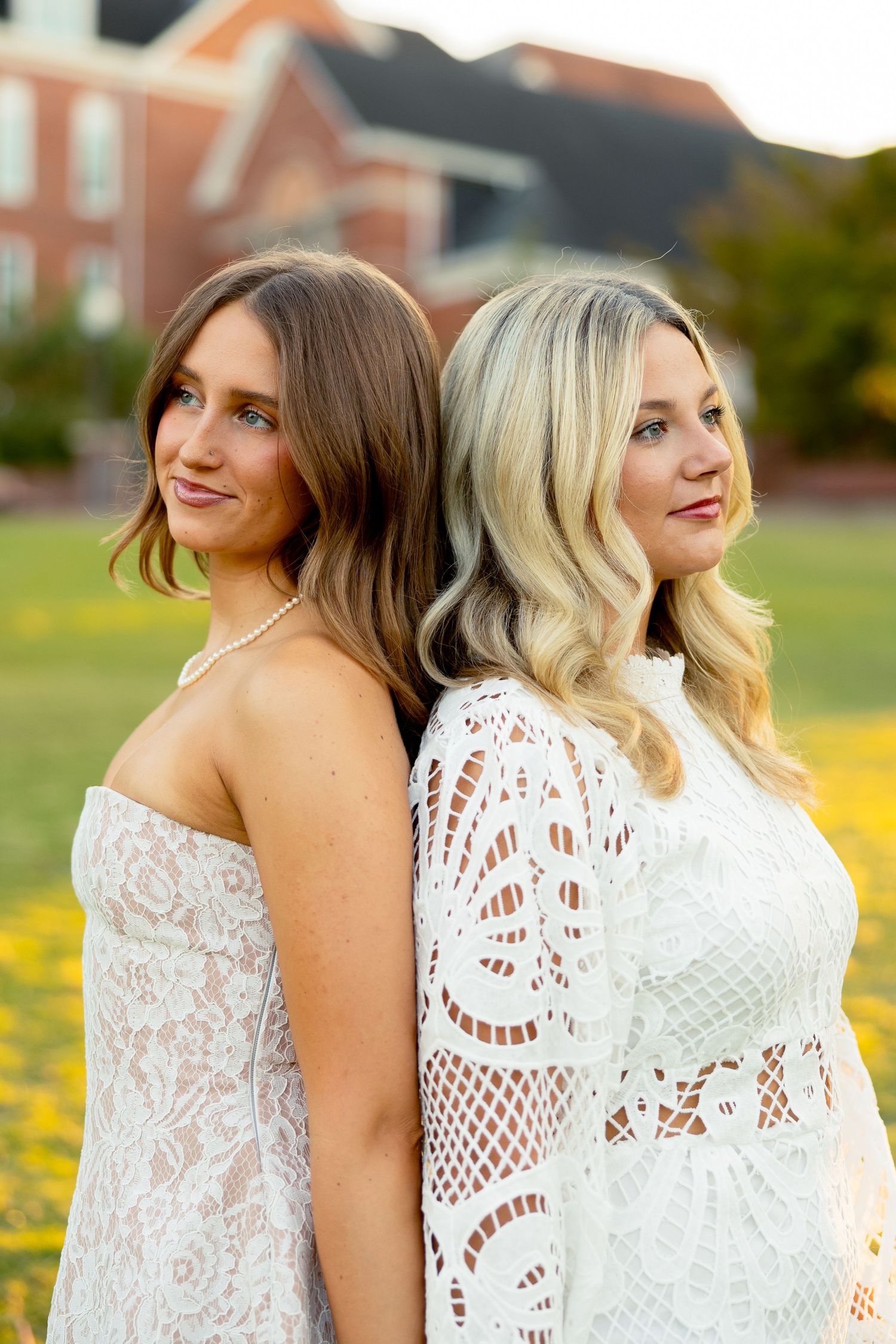 Two young women in white lace dresses stand back-to-back in a sunny field, with a brick building in the background.