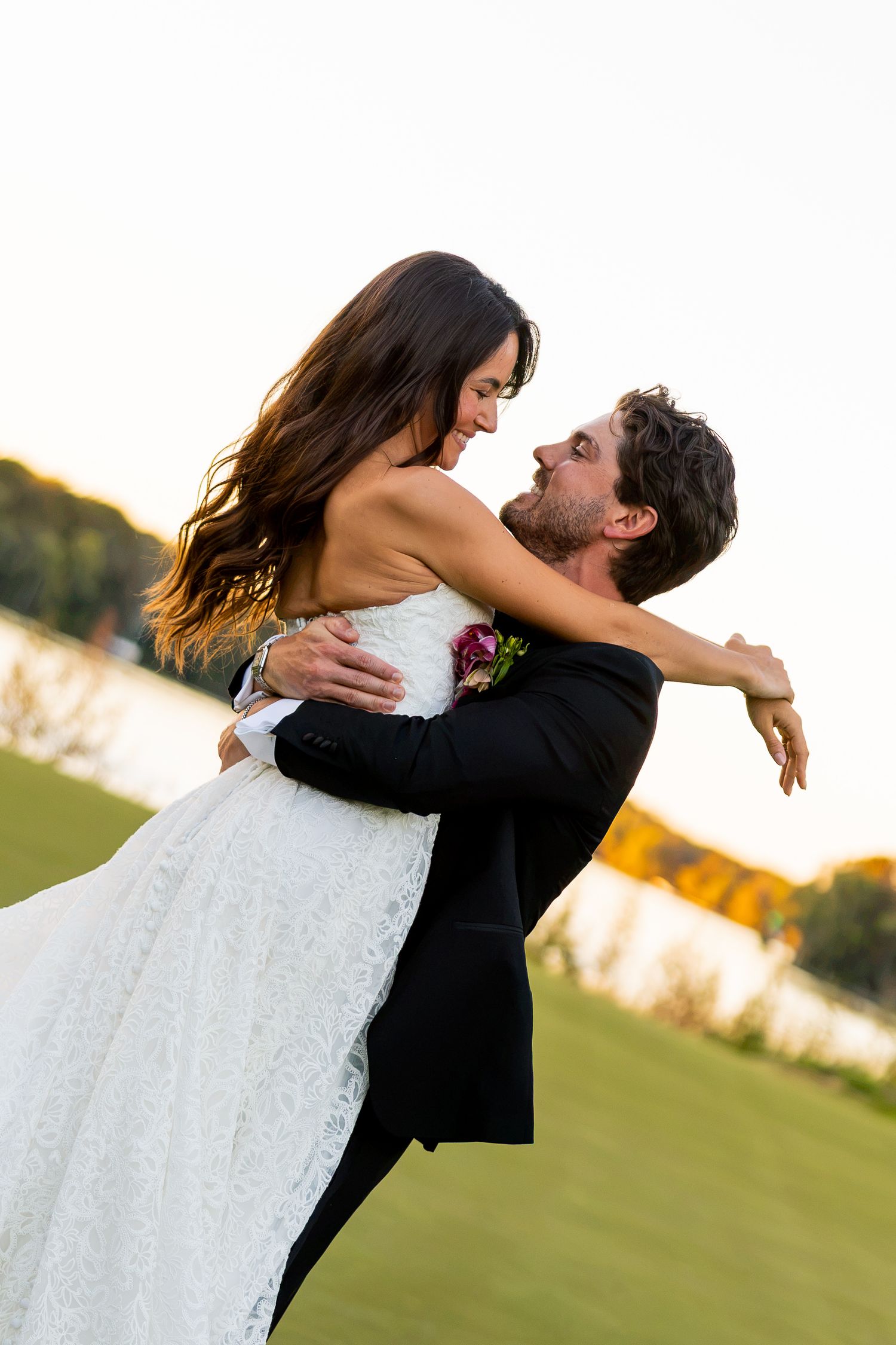 Groom lifts bride in white lace gown outdoors at golden hour, both smiling joyfully near a serene lakeside setting.