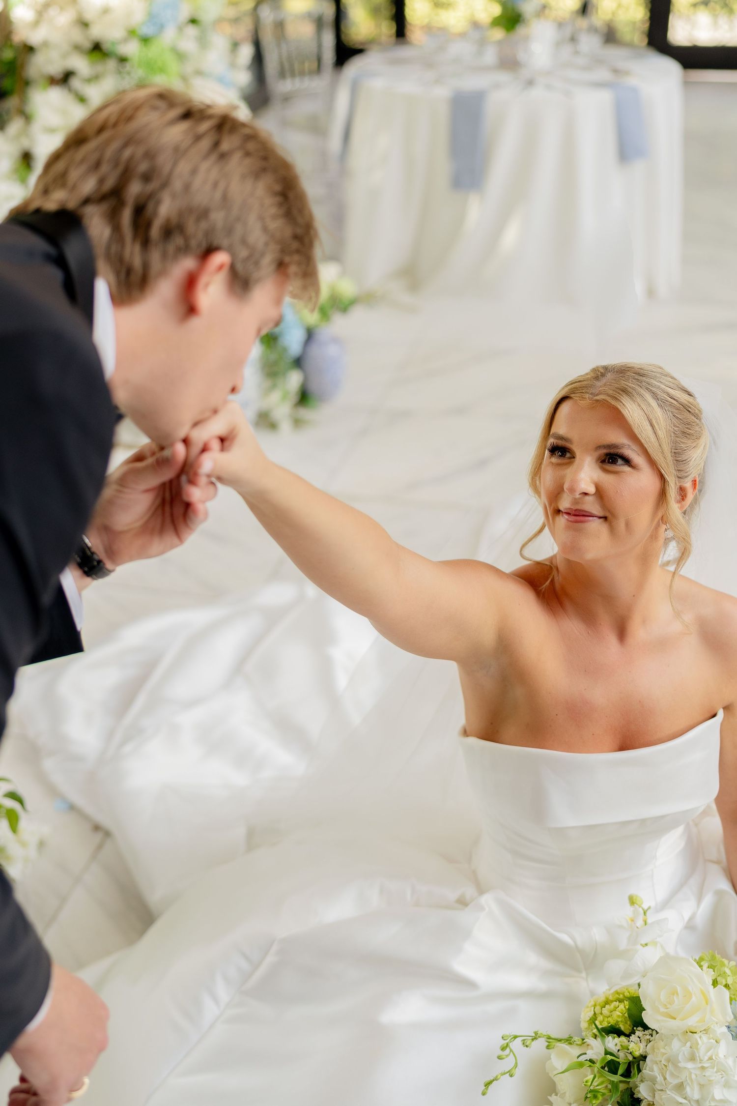 Bride in white strapless gown extends hand for groom to kiss at elegant floral wedding reception.