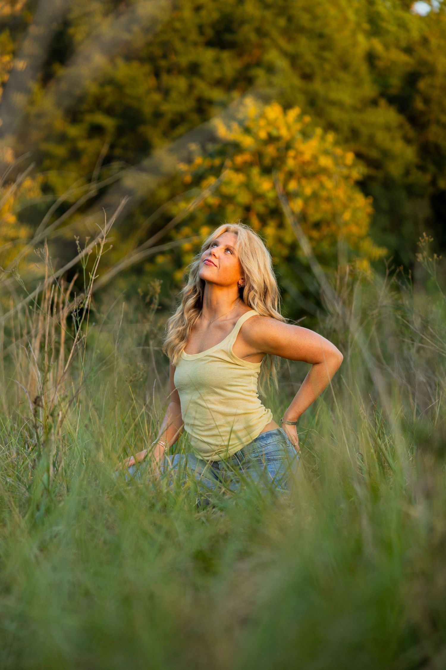 Blonde woman in yellow tank top sitting in tall green grass, looking up, surrounded by autumn trees in golden light.