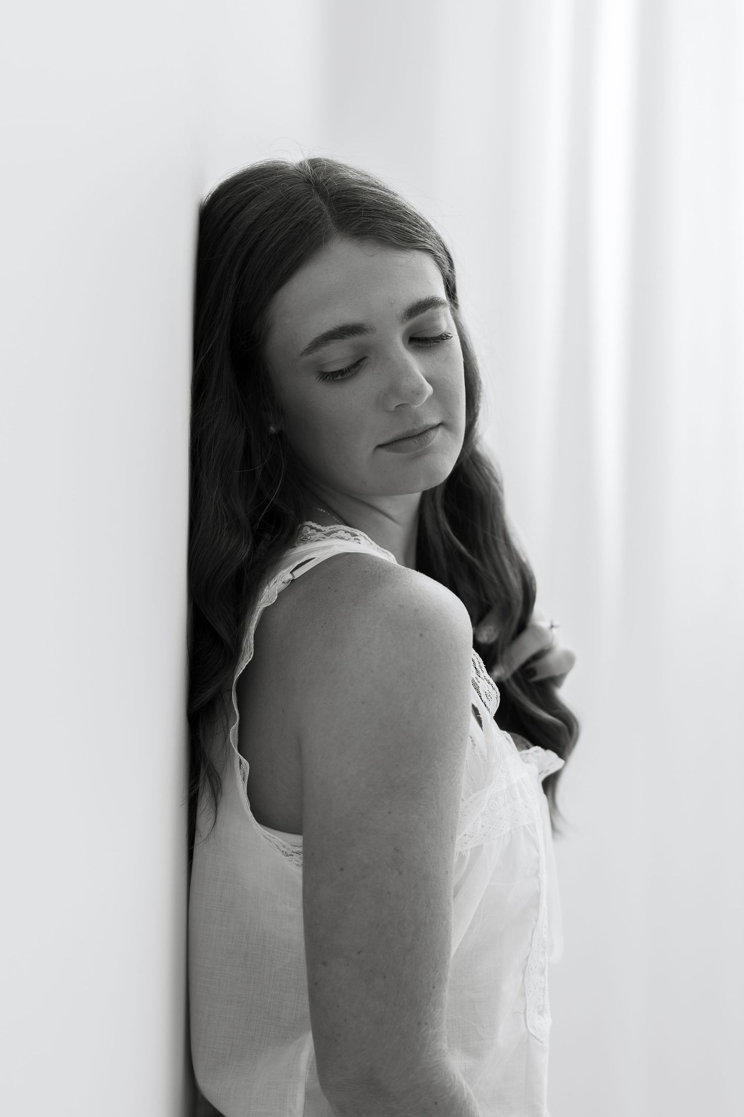 Black and white portrait of a young woman with long hair leaning against a white wall, eyes closed, wearing a white dress.