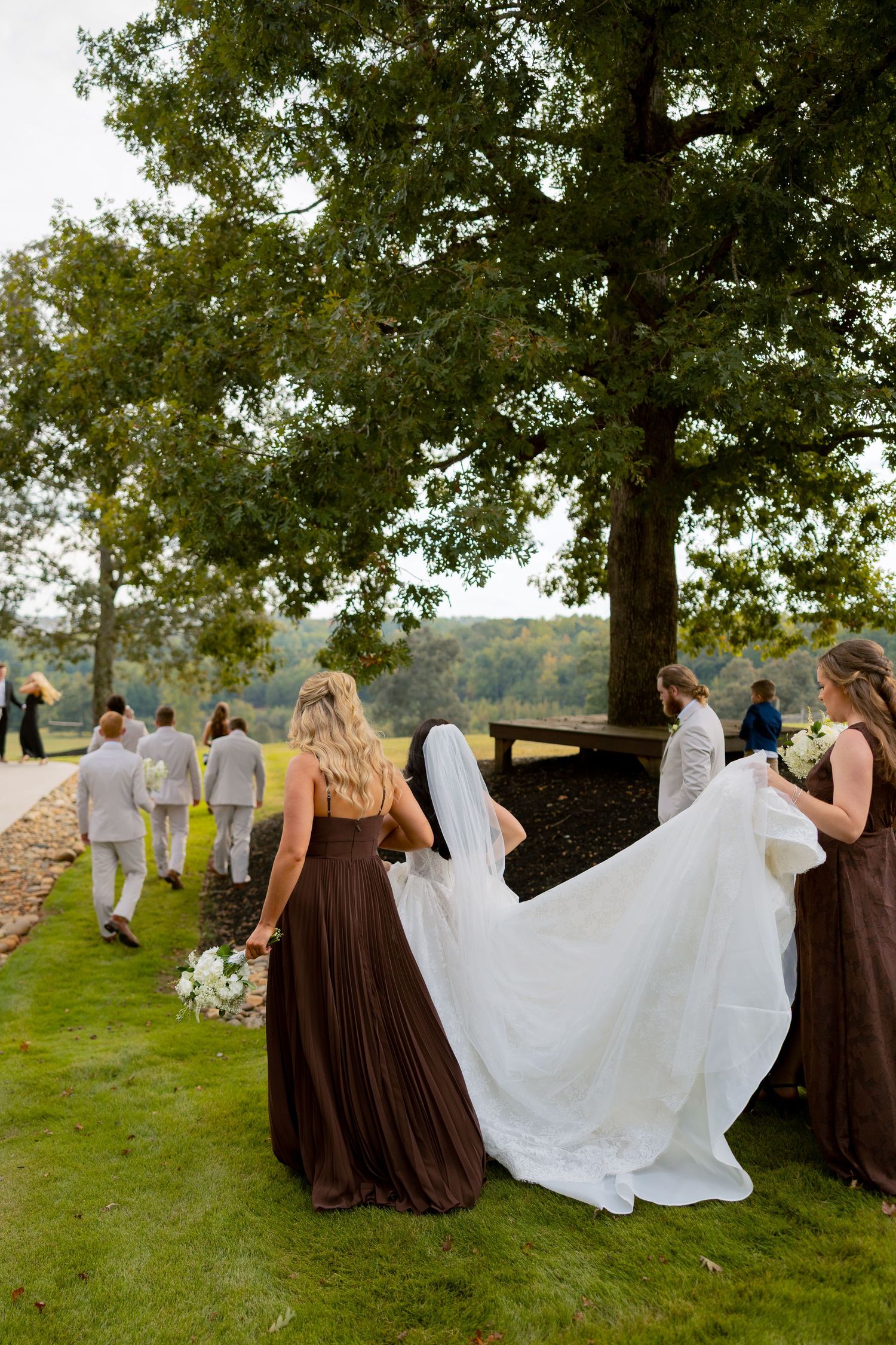Bridesmaids in brown dresses help carry a bride's white train outdoors near large trees at a wedding.