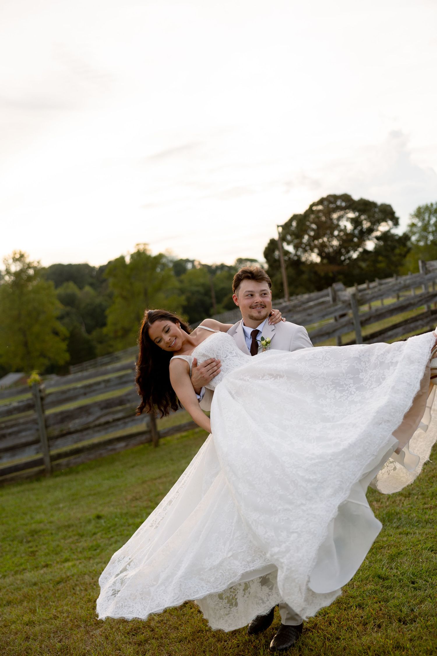 Groom carrying bride in white lace wedding dress outdoors on green grass with rustic fence and trees in background.