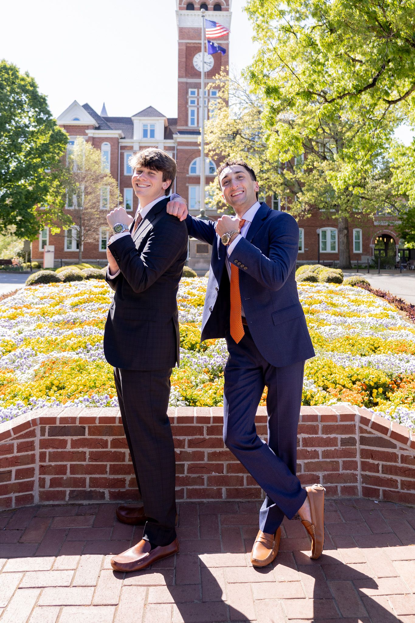 Two young men in suits pose confidently outdoors near a brick university building with colorful flower beds.