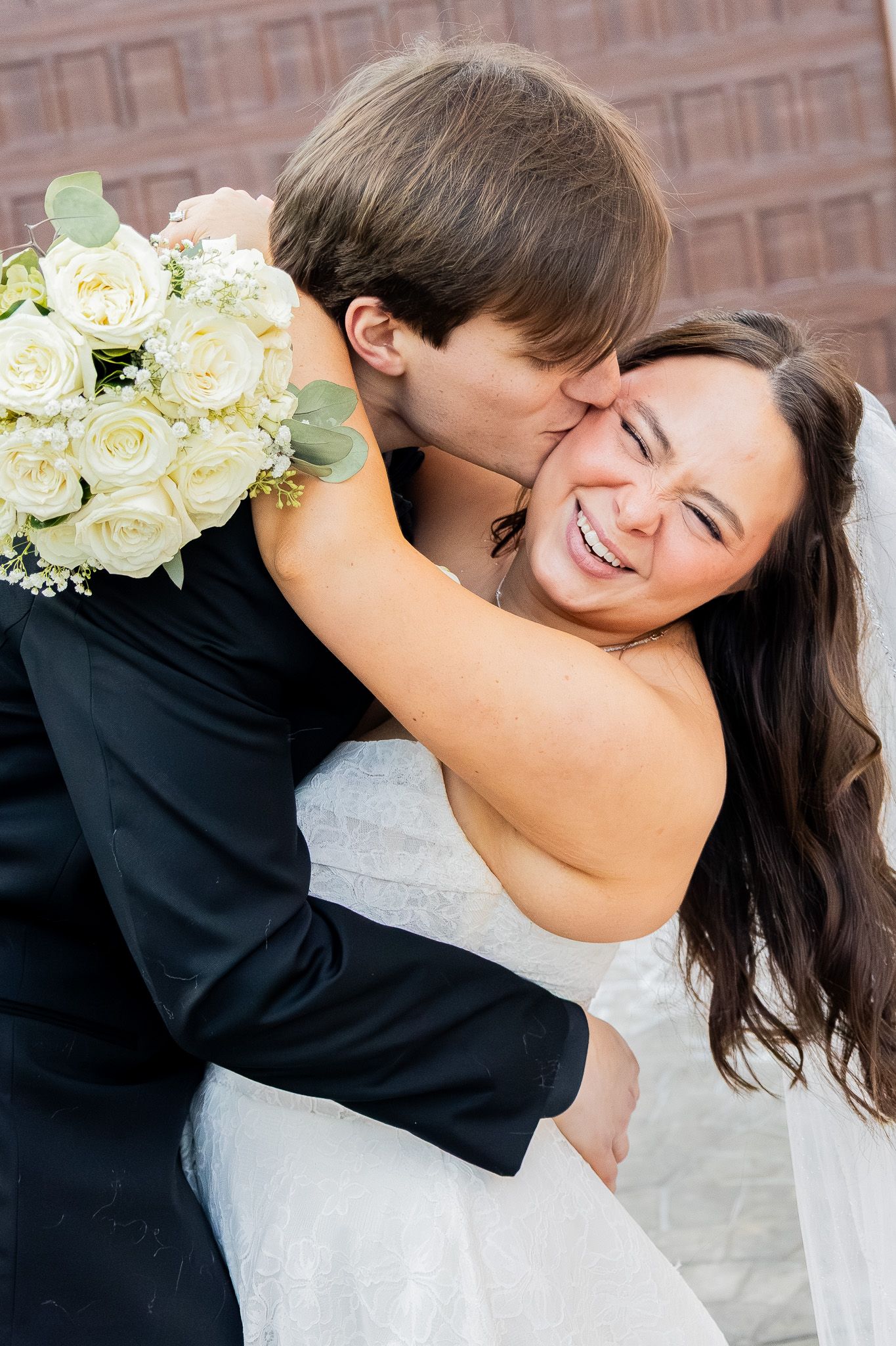 Joyful bride in white dress laughs as groom in black tuxedo kisses her cheek while holding rose bouquet.