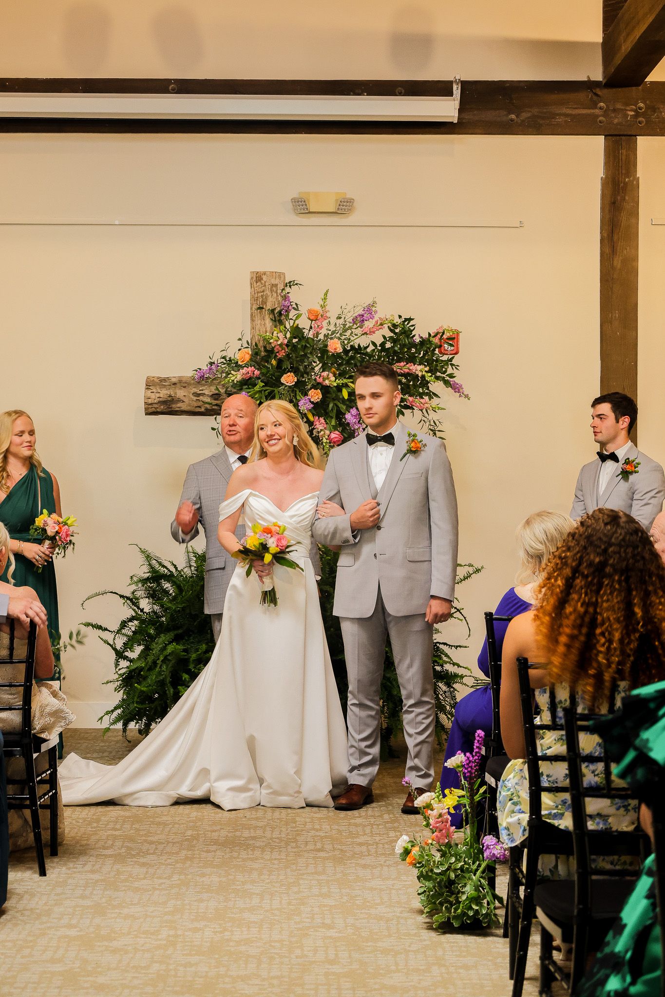 Bride and groom stand at altar with floral cross backdrop during indoor wedding ceremony, surrounded by guests.