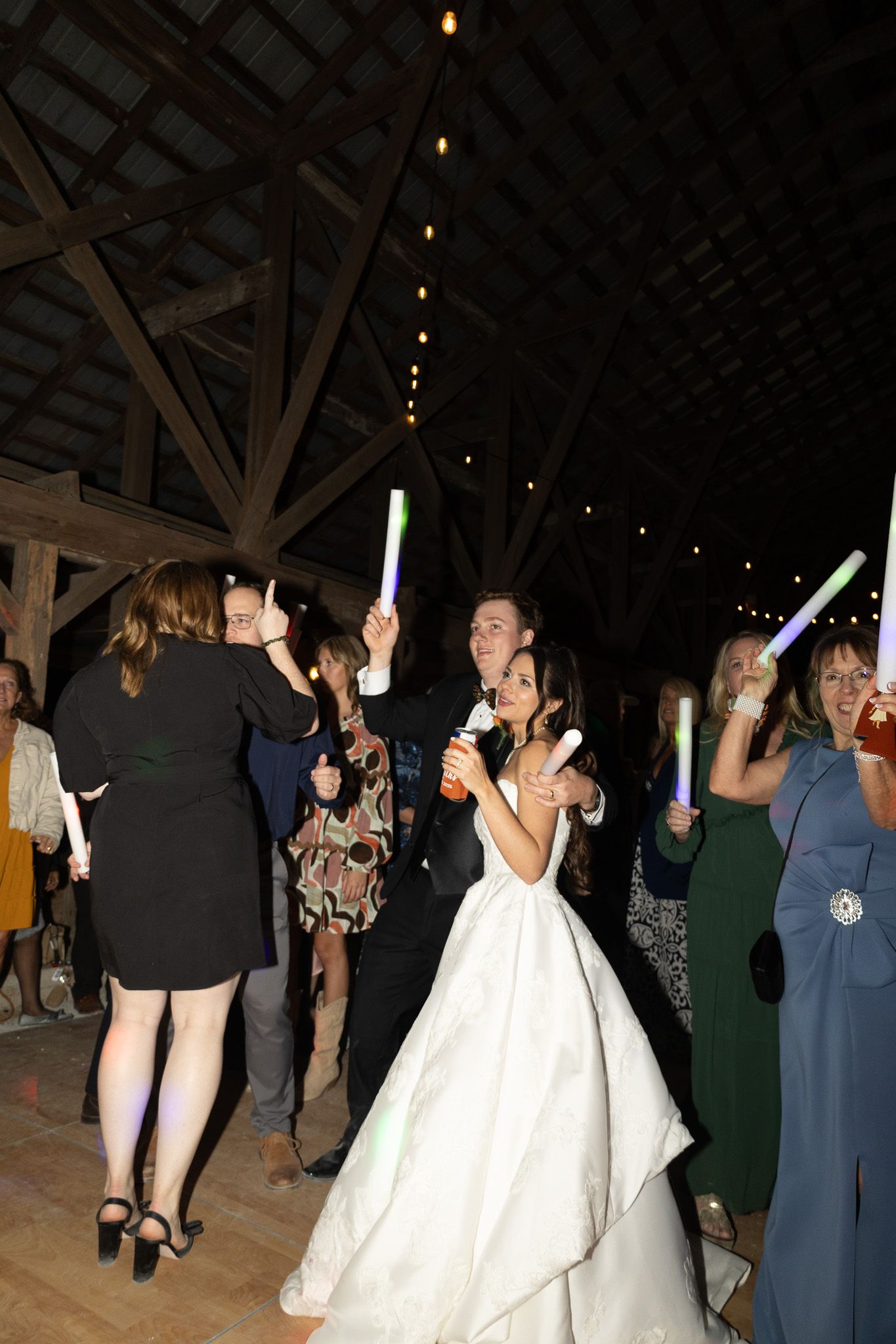 Bride and groom dancing with glowing foam sticks at rustic barn wedding reception with string lights.