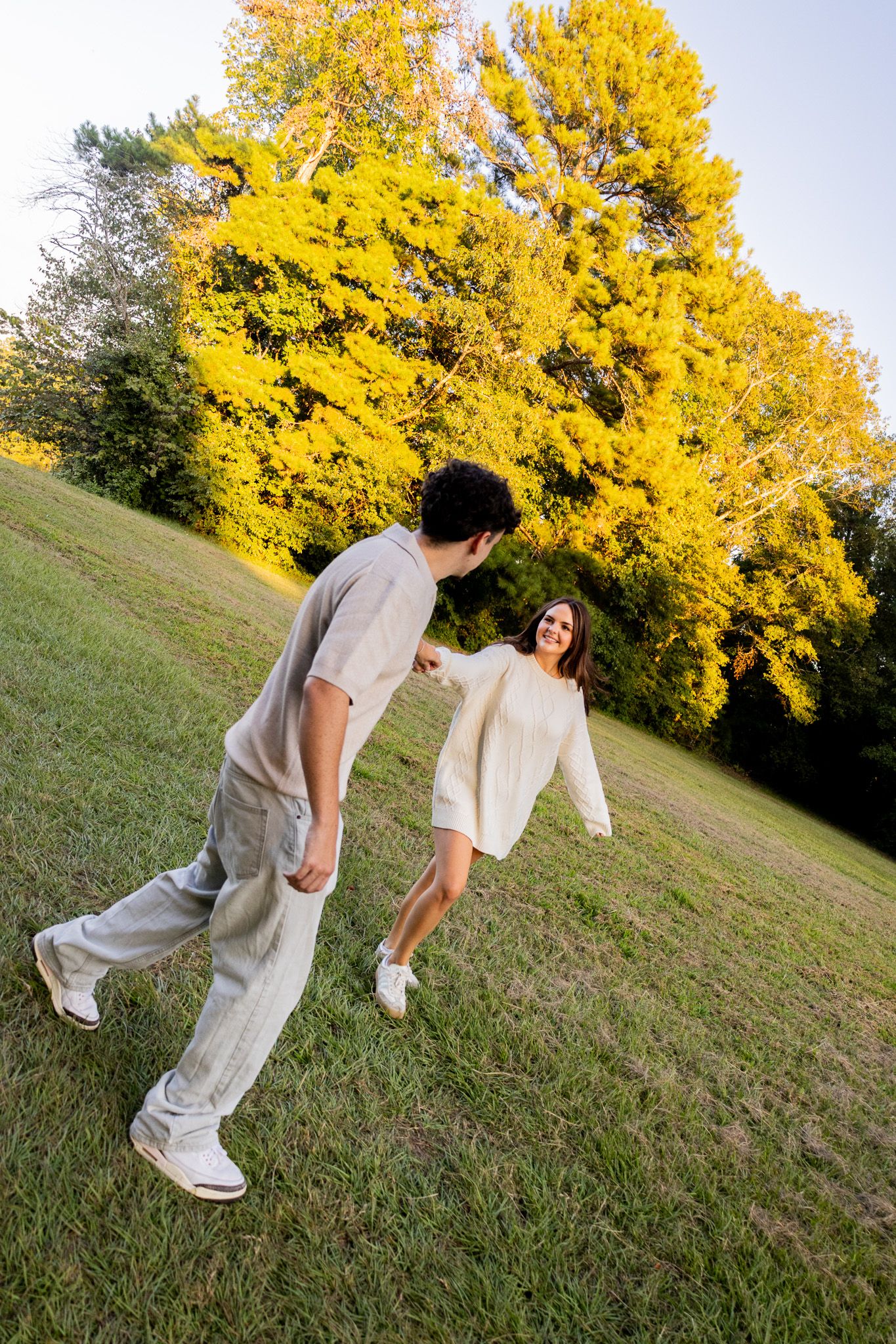 A couple holds hands and plays on a grassy hill surrounded by vibrant yellow autumn trees at golden hour.