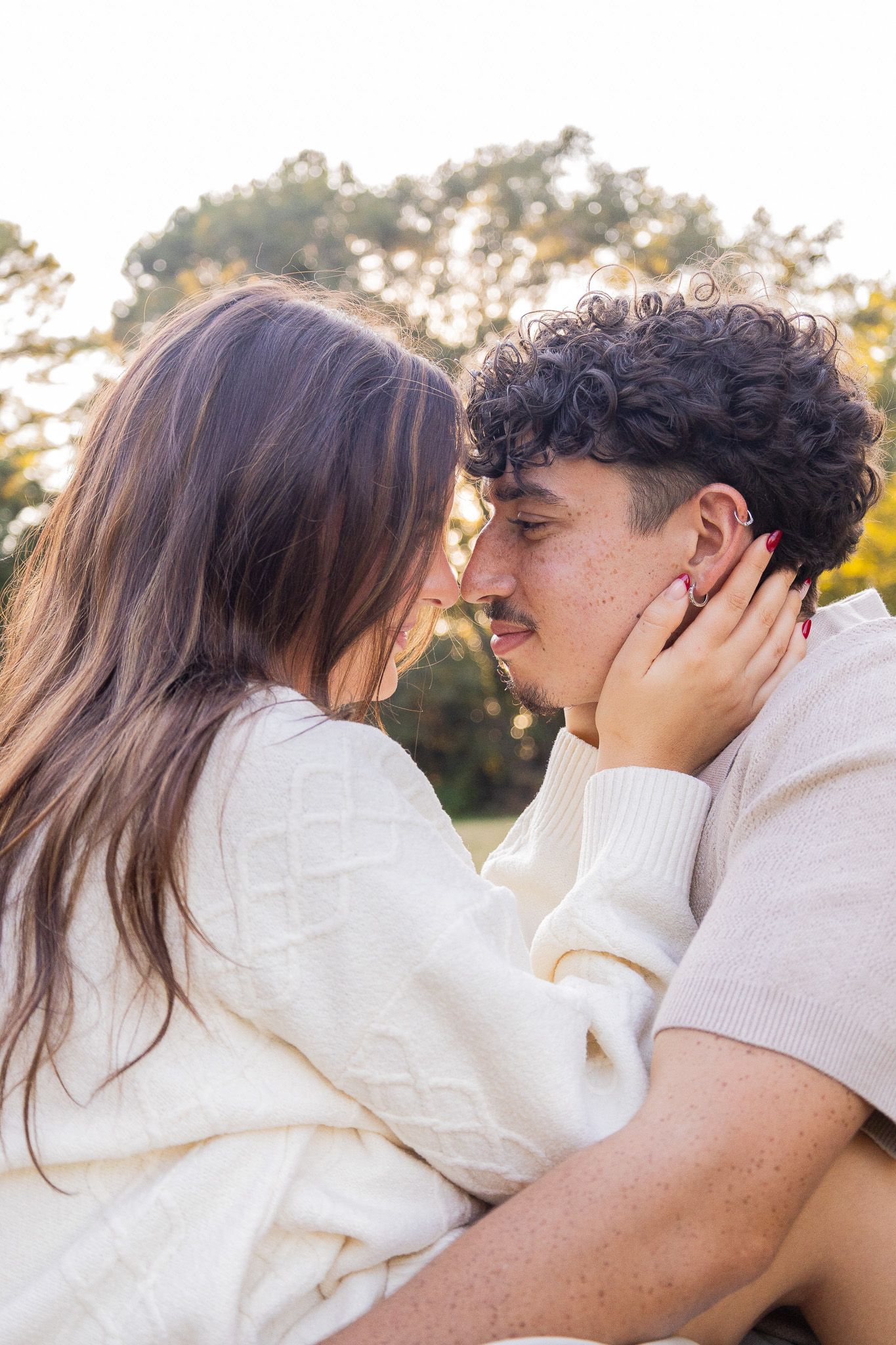 A young couple in matching cream sweaters share a tender romantic moment outdoors, with the woman gently cradling the man's face.
