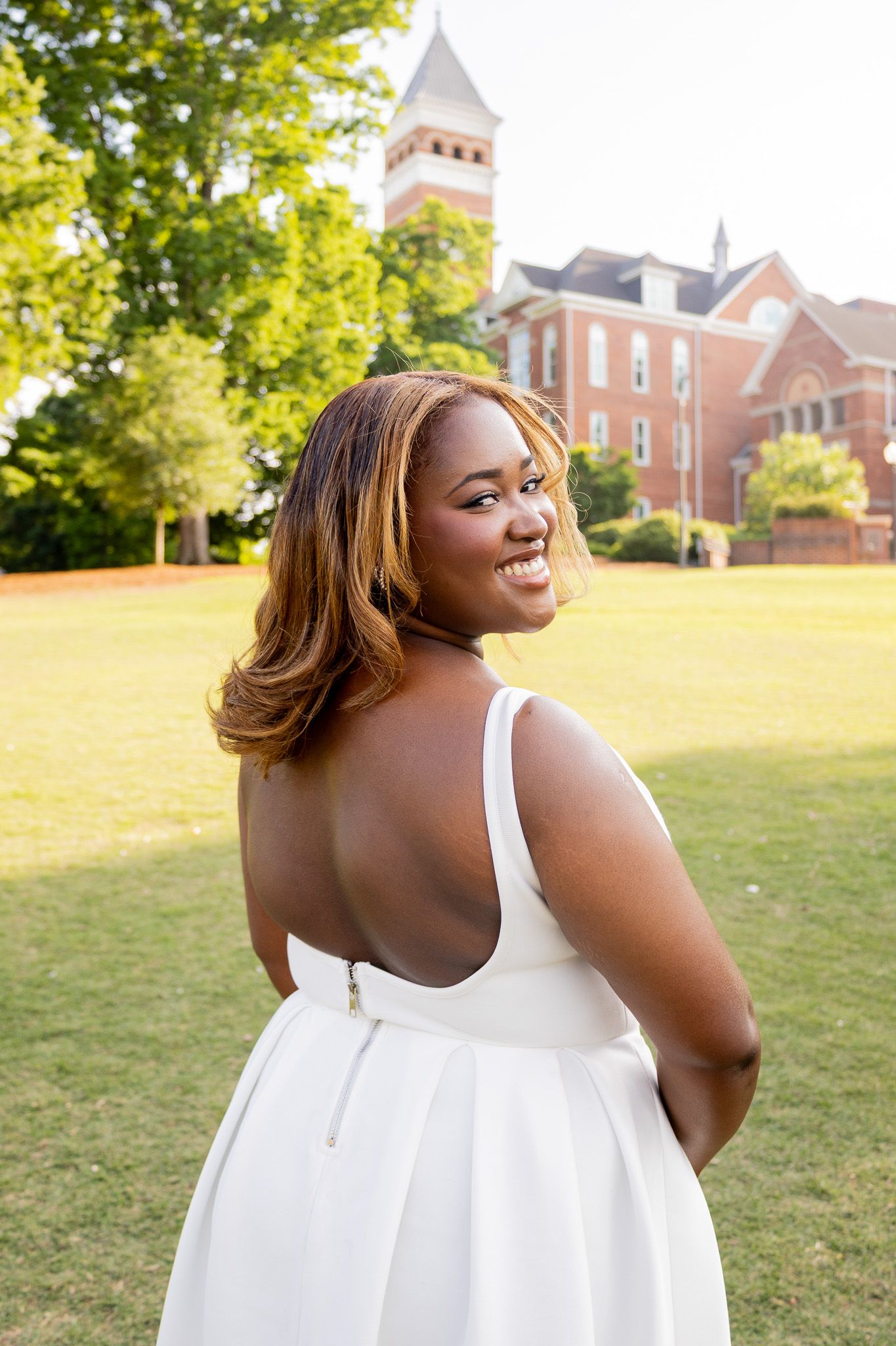 A smiling young Black woman in a white backless dress poses outdoors near a red brick building on a sunny day.