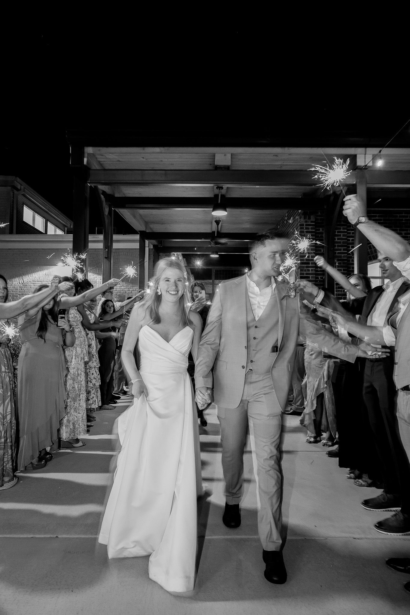 Black and white photo of a smiling bride and groom walking through a sparkler send-off at their wedding reception.