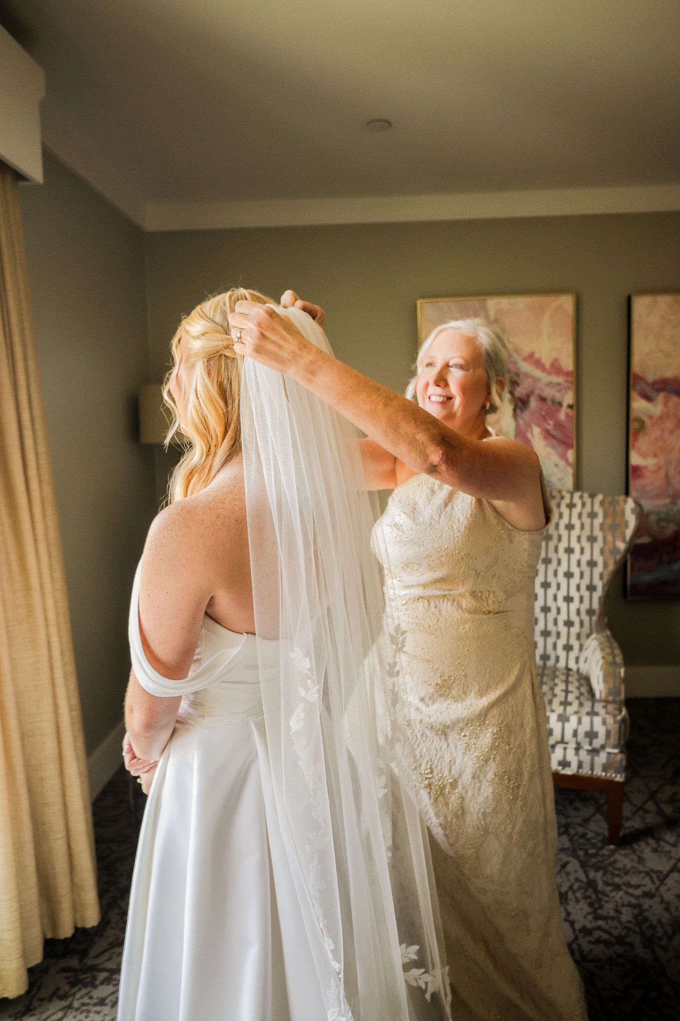 Mother helping bride put on wedding veil in a hotel room before the ceremony.