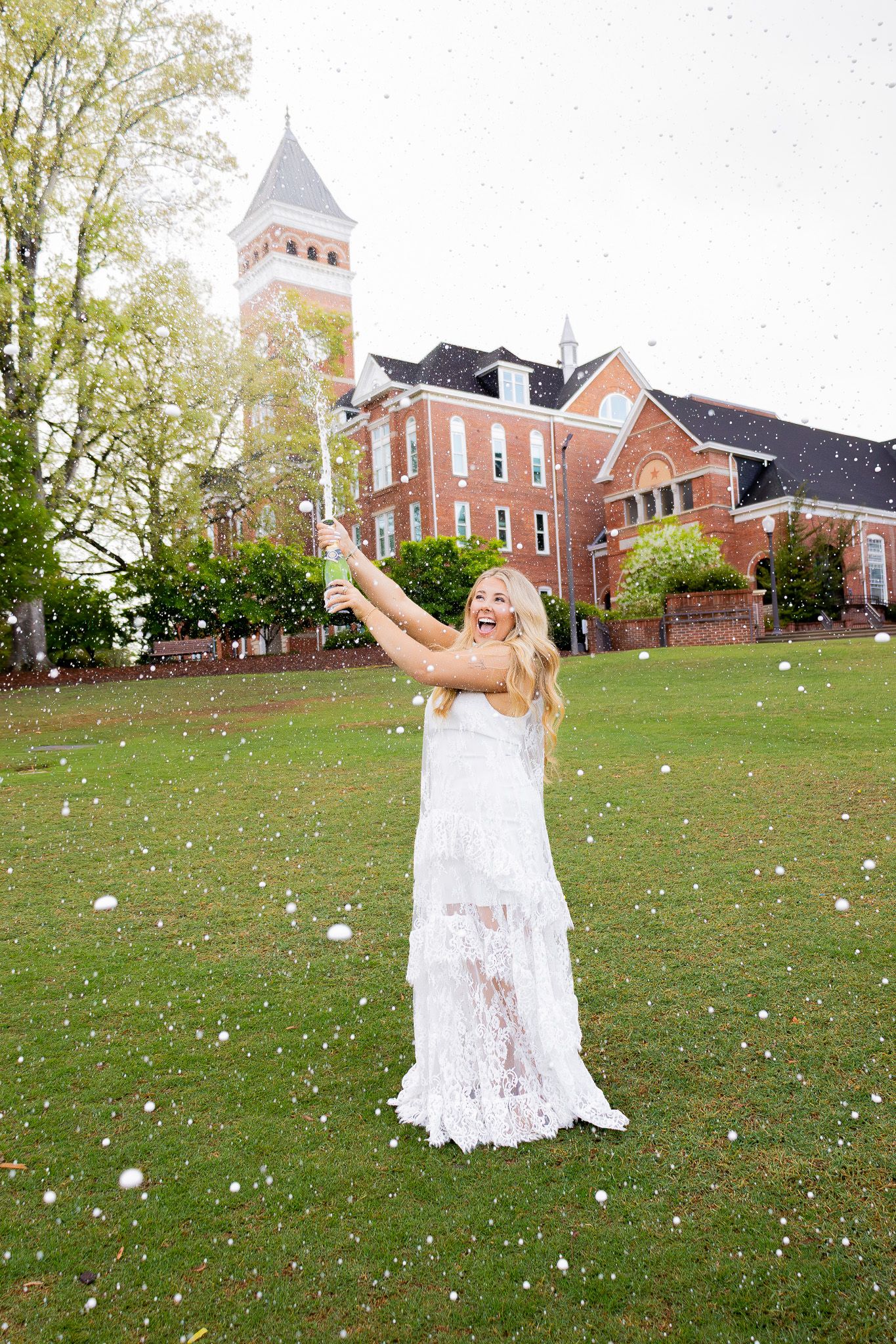 Excited blonde woman in white lace dress celebrating outdoors on Clemson University campus lawn with confetti.