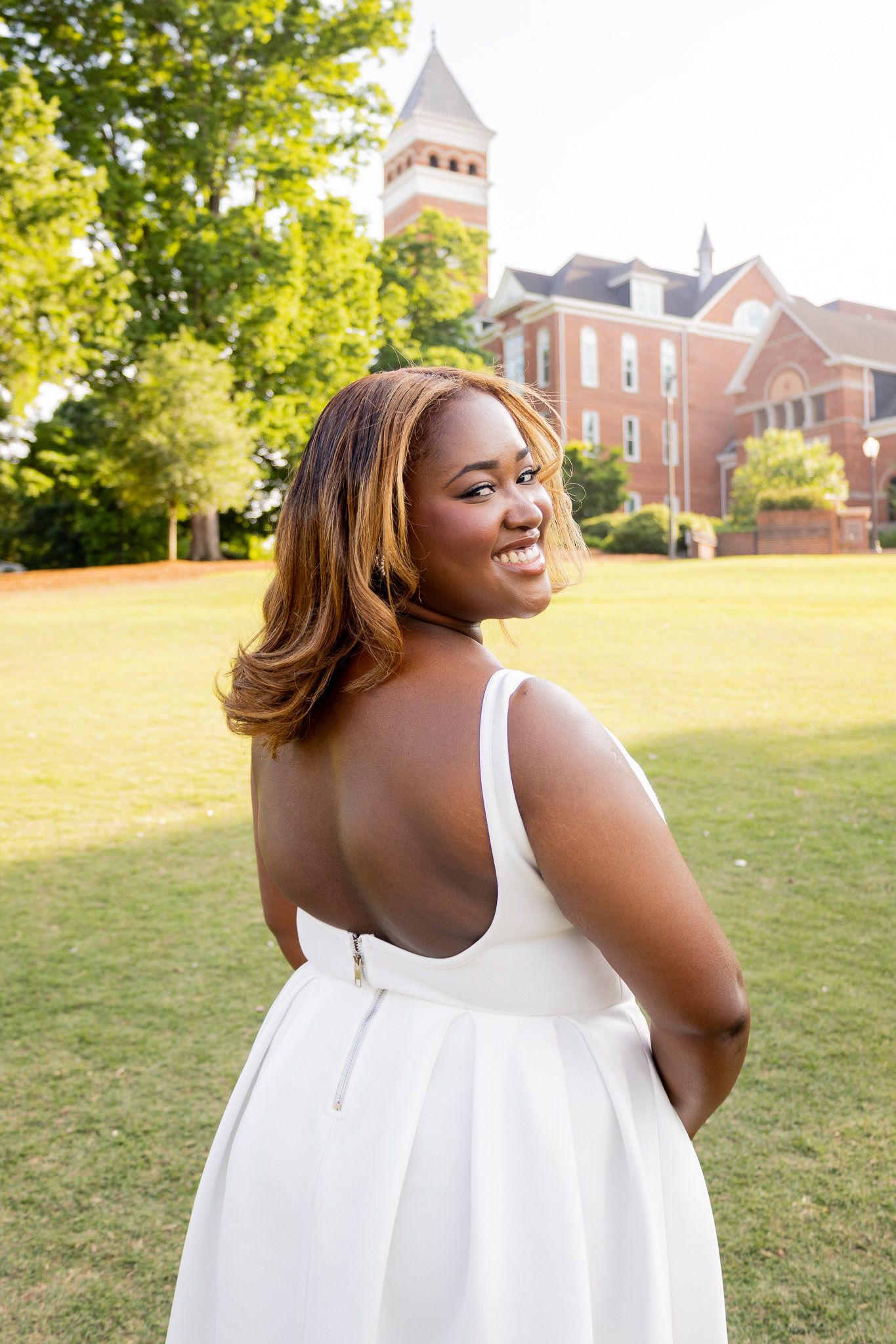 Young woman in white backless dress smiles over shoulder on green campus lawn with brick building backdrop.