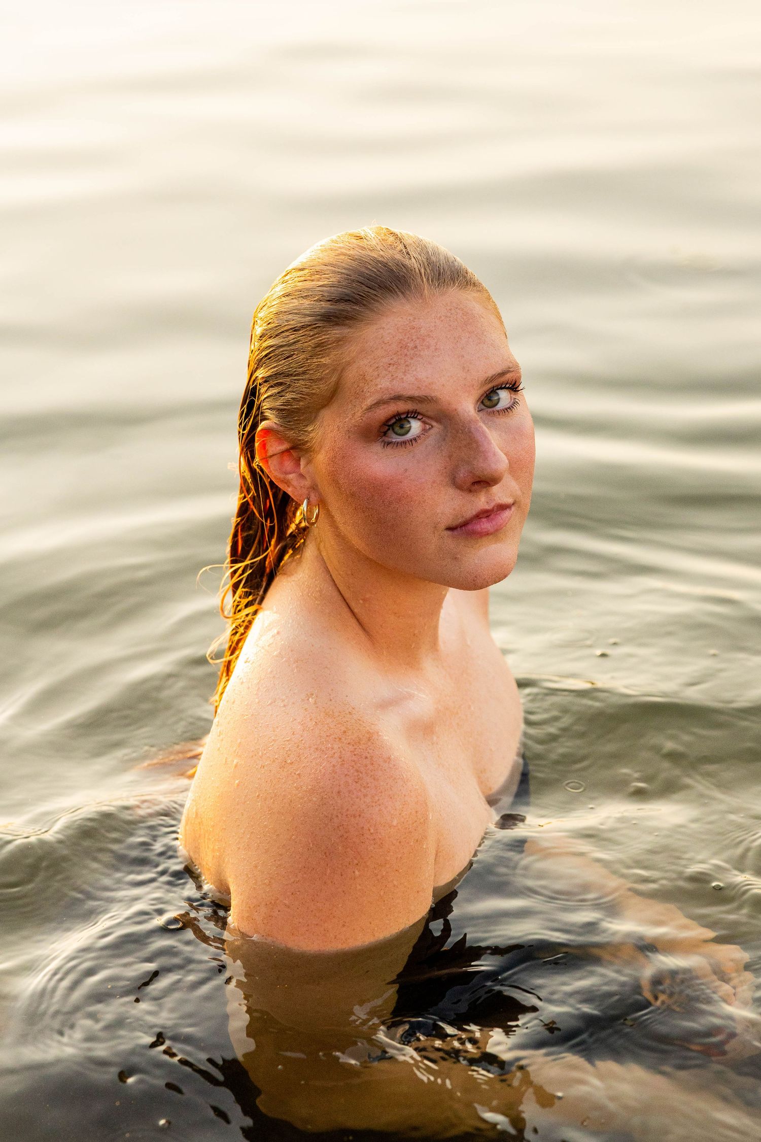 Young woman with wet hair and freckles emerging from calm golden water at sunset.