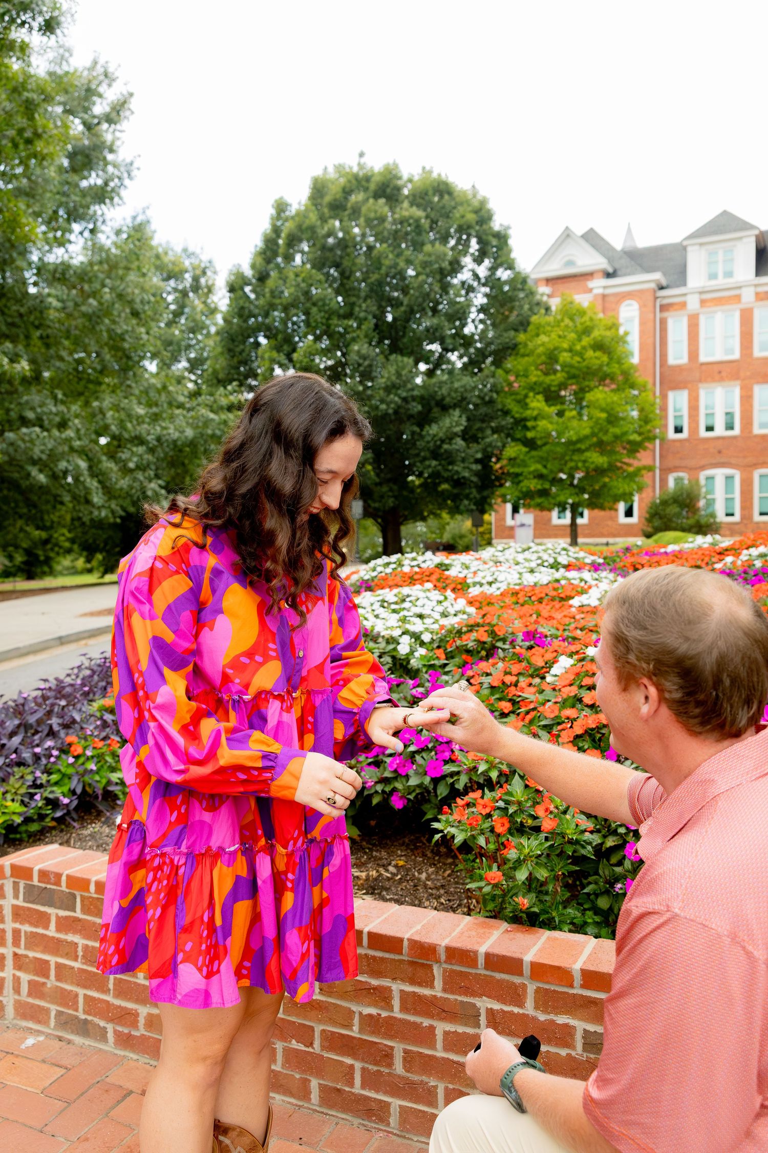 Man kneeling to propose to woman in colorful pink dress beside brick flower garden with red building backdrop.