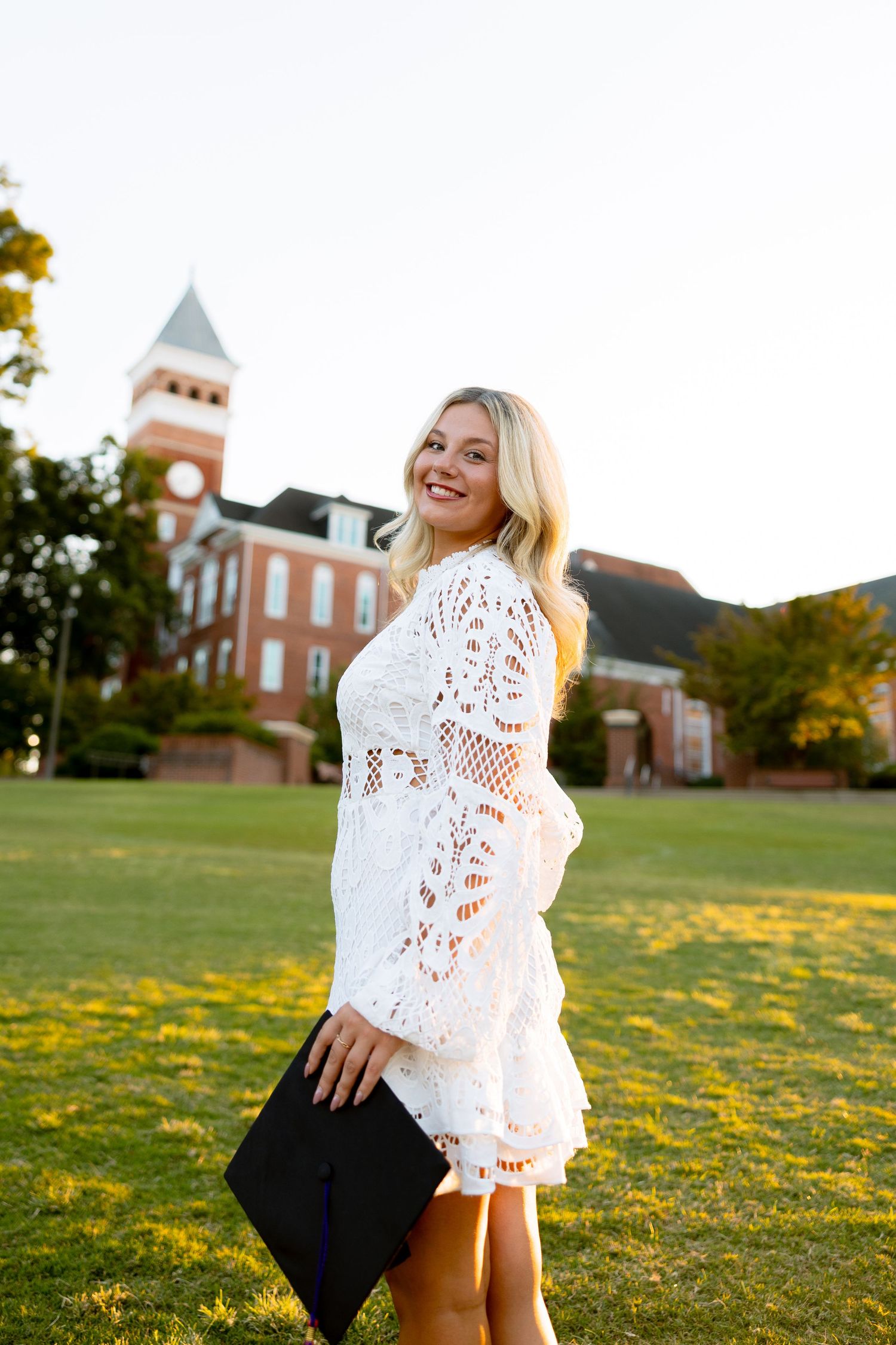 Smiling blonde woman in white lace dress holding graduation cap on green campus lawn with brick building backdrop.