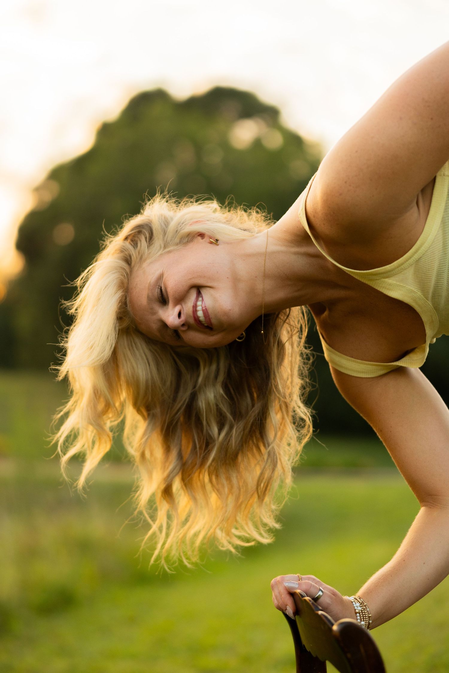 Smiling blonde woman bending over outdoors at sunset in a park, hair flowing down, wearing a yellow top.