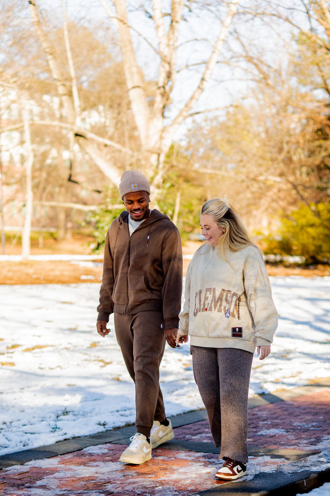 Couple walking outdoors in winter wearing Clemson University sweatshirt and brown tracksuit on snowy path.