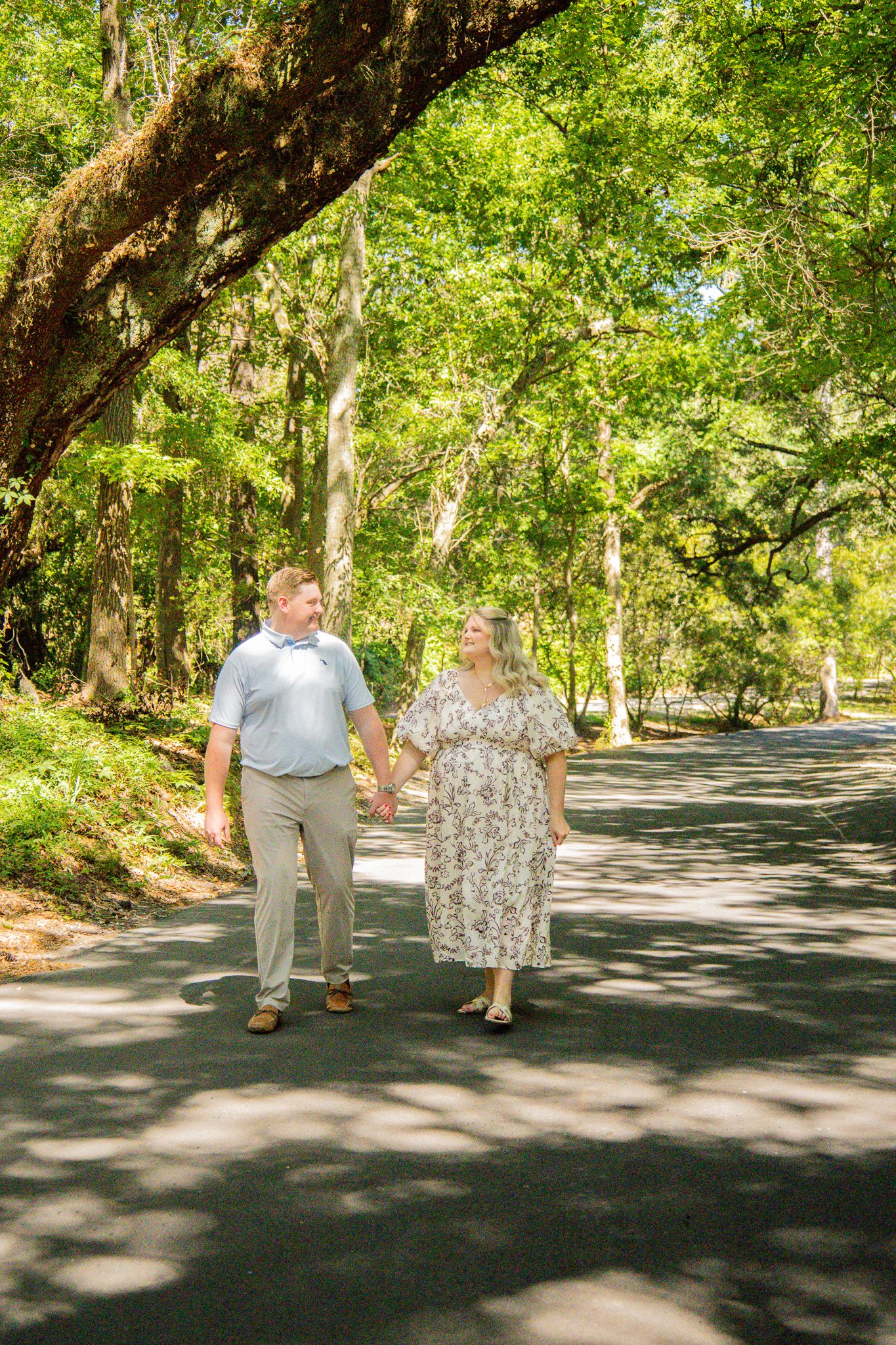 A couple holds hands walking down a tree-lined path surrounded by lush green foliage on a sunny day.