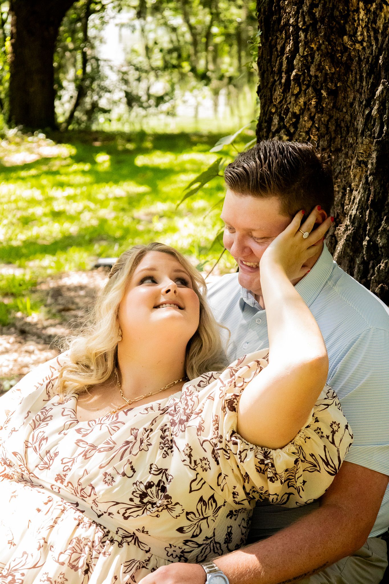 A couple shares a tender moment leaning against a tree in a sunny park, the woman gazing up lovingly at the smiling man.