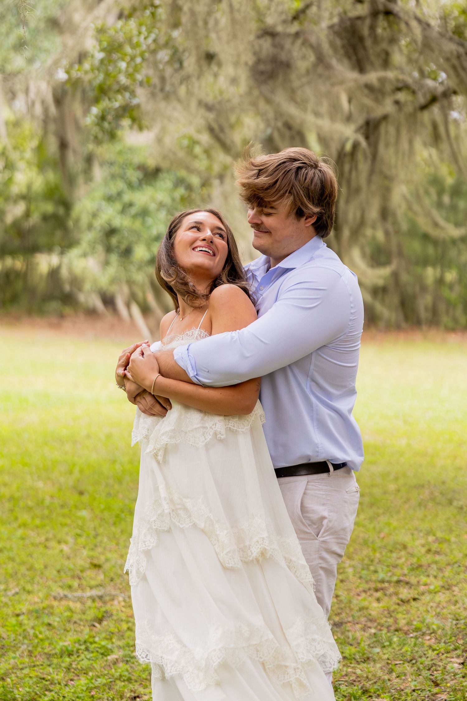 A smiling woman in a white lace dress looks up as a man in a blue shirt hugs her from behind on a grassy lawn beneath moss-draped oak trees.