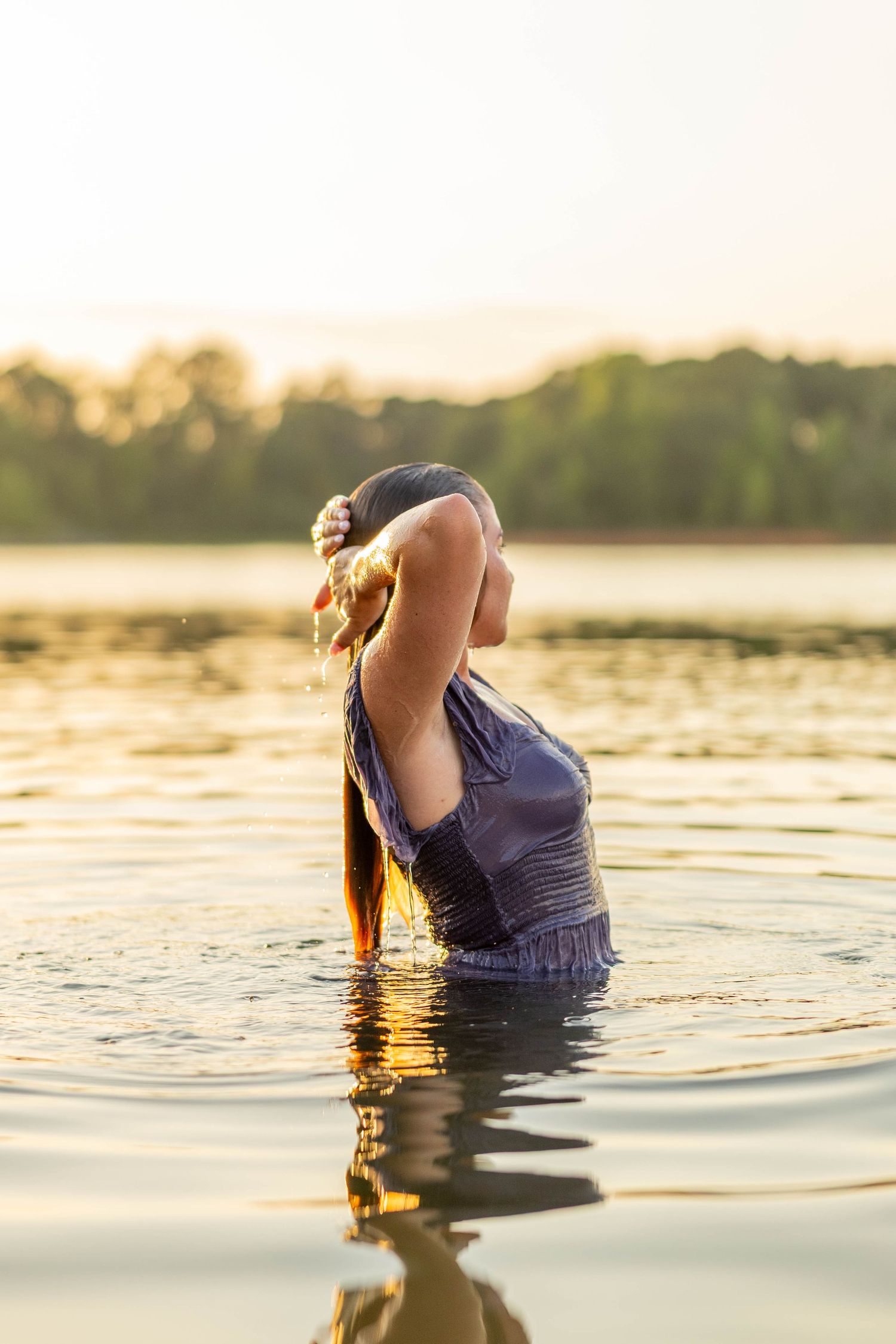 Woman standing in a lake at golden hour, wearing a blue dress with wet hair, surrounded by calm water.