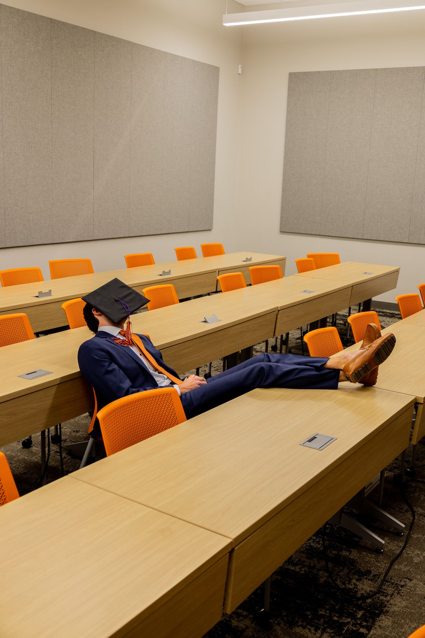Graduate in cap and suit relaxing in empty classroom with orange chairs and wooden desks after graduation.