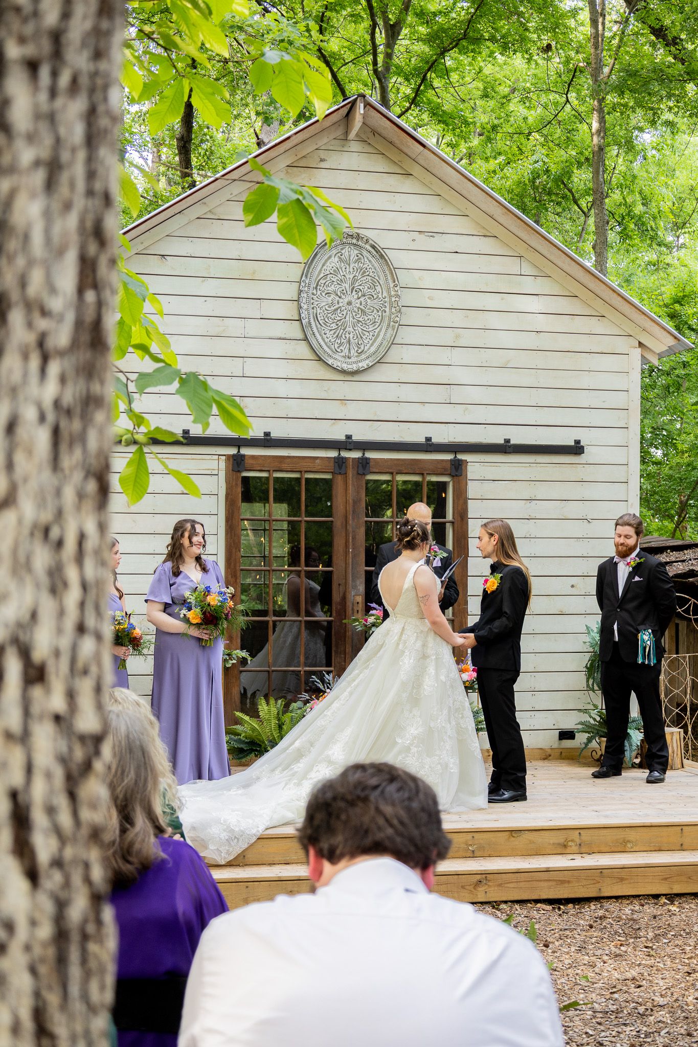 Outdoor wedding ceremony in front of a white chapel with barn doors, bride in lace gown, bridesmaid in lavender dress.