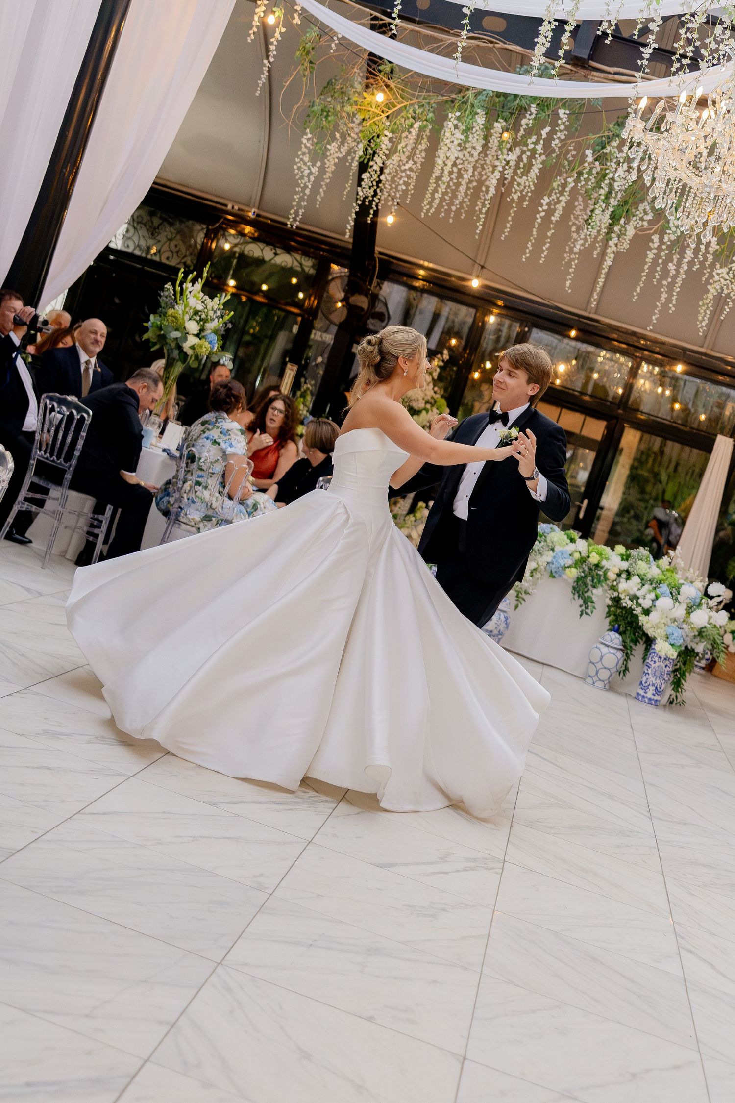 Bride and groom share their first dance on a white marble floor at an elegant wedding reception with floral decor.