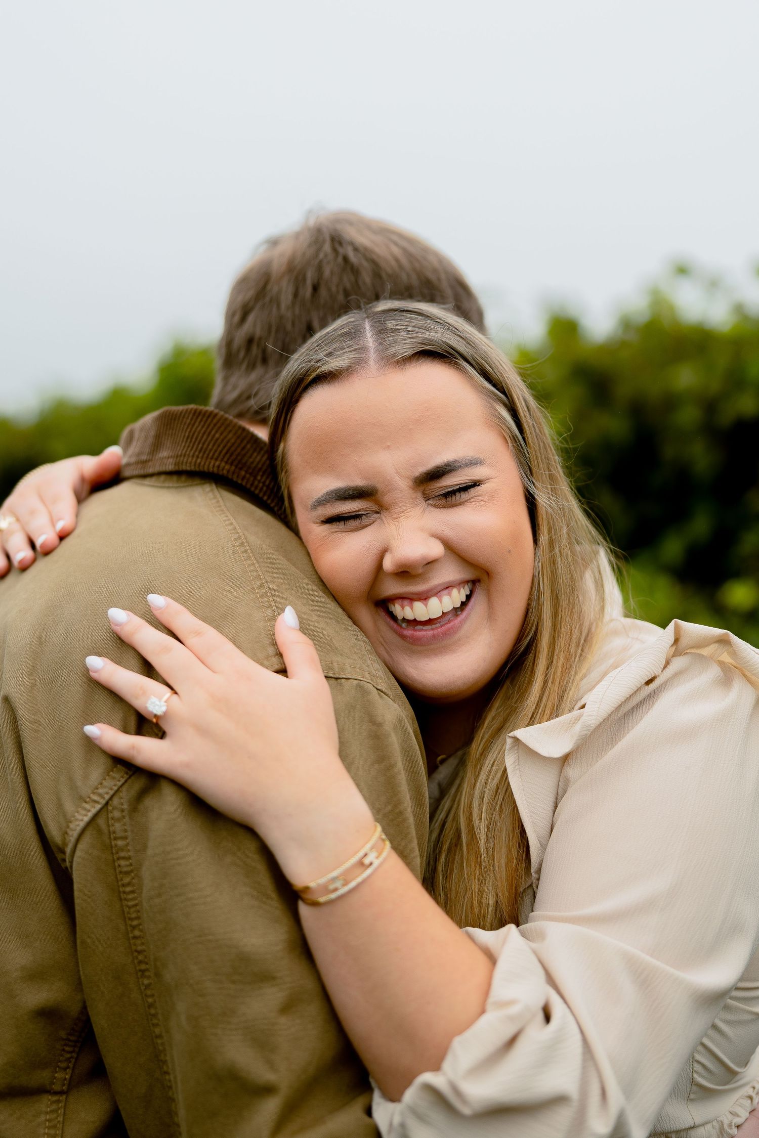 A joyful woman laughs while hugging a man outdoors, showcasing her engagement ring and bracelet.