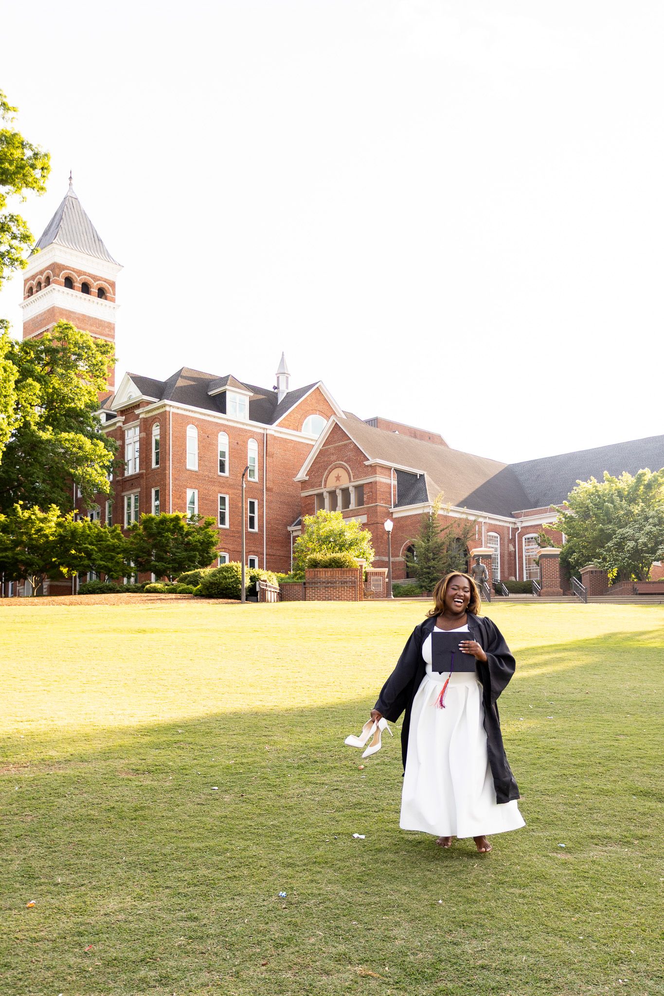 Graduate in cap and gown stands on green campus lawn before historic red brick university building.