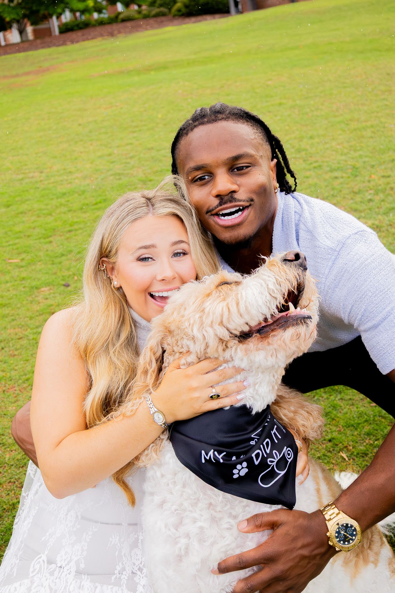 Couple smiling and hugging a fluffy golden dog wearing a bandana on a green lawn.