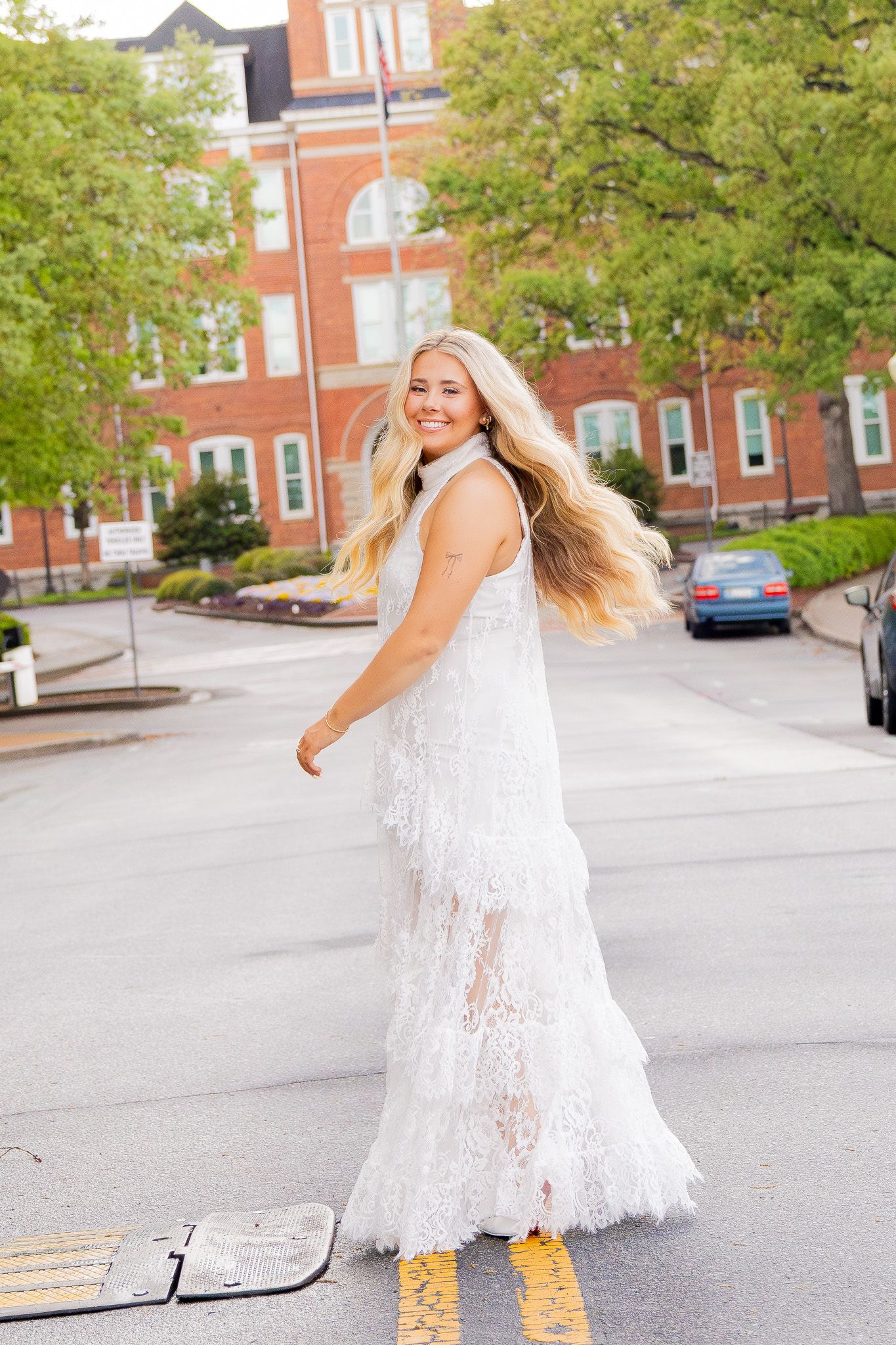 Blonde woman twirling in white lace fringe maxi dress on urban street with brick building backdrop.