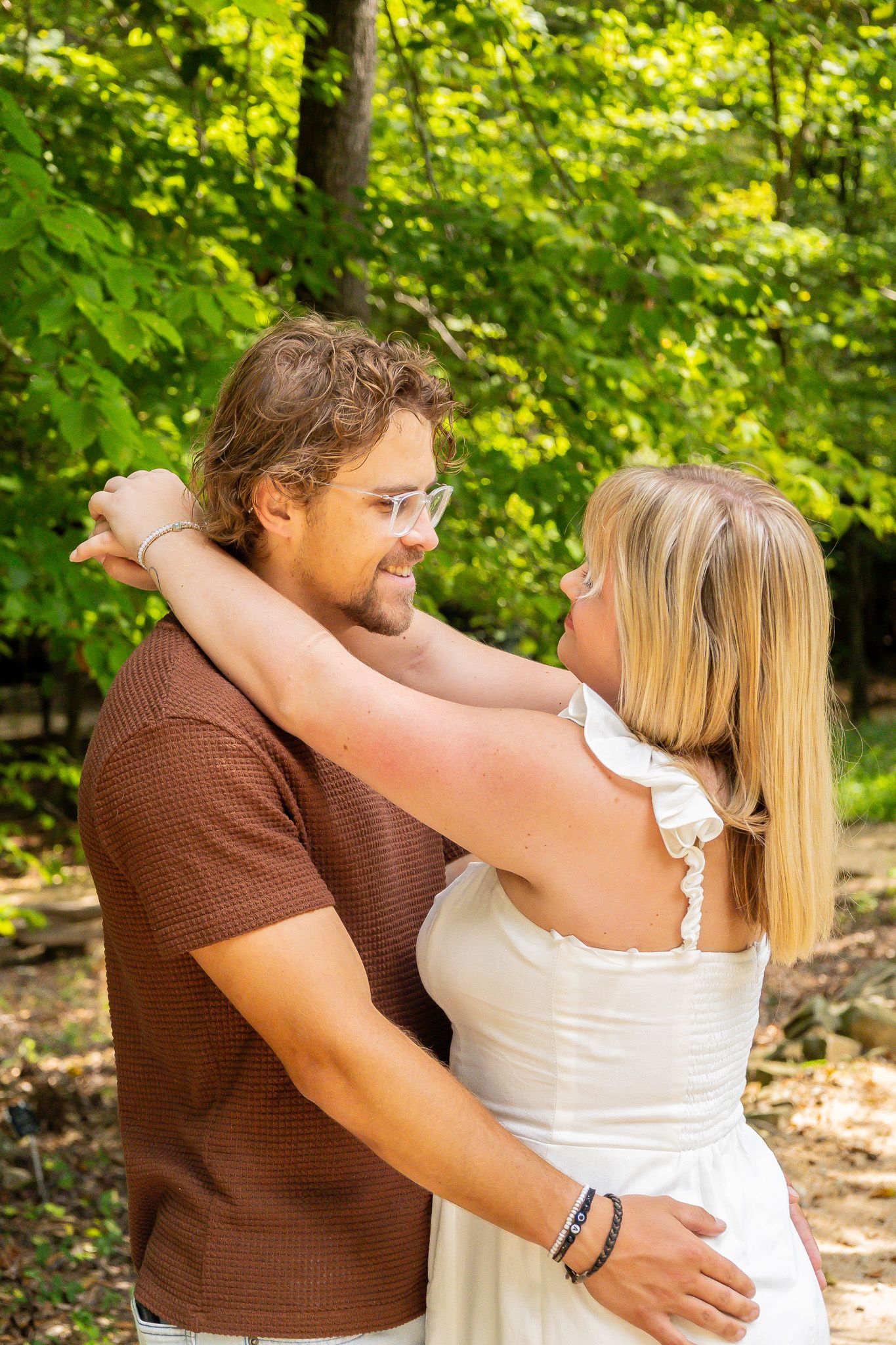 A couple embraces outdoors, smiling at each other surrounded by lush green trees in a romantic setting.