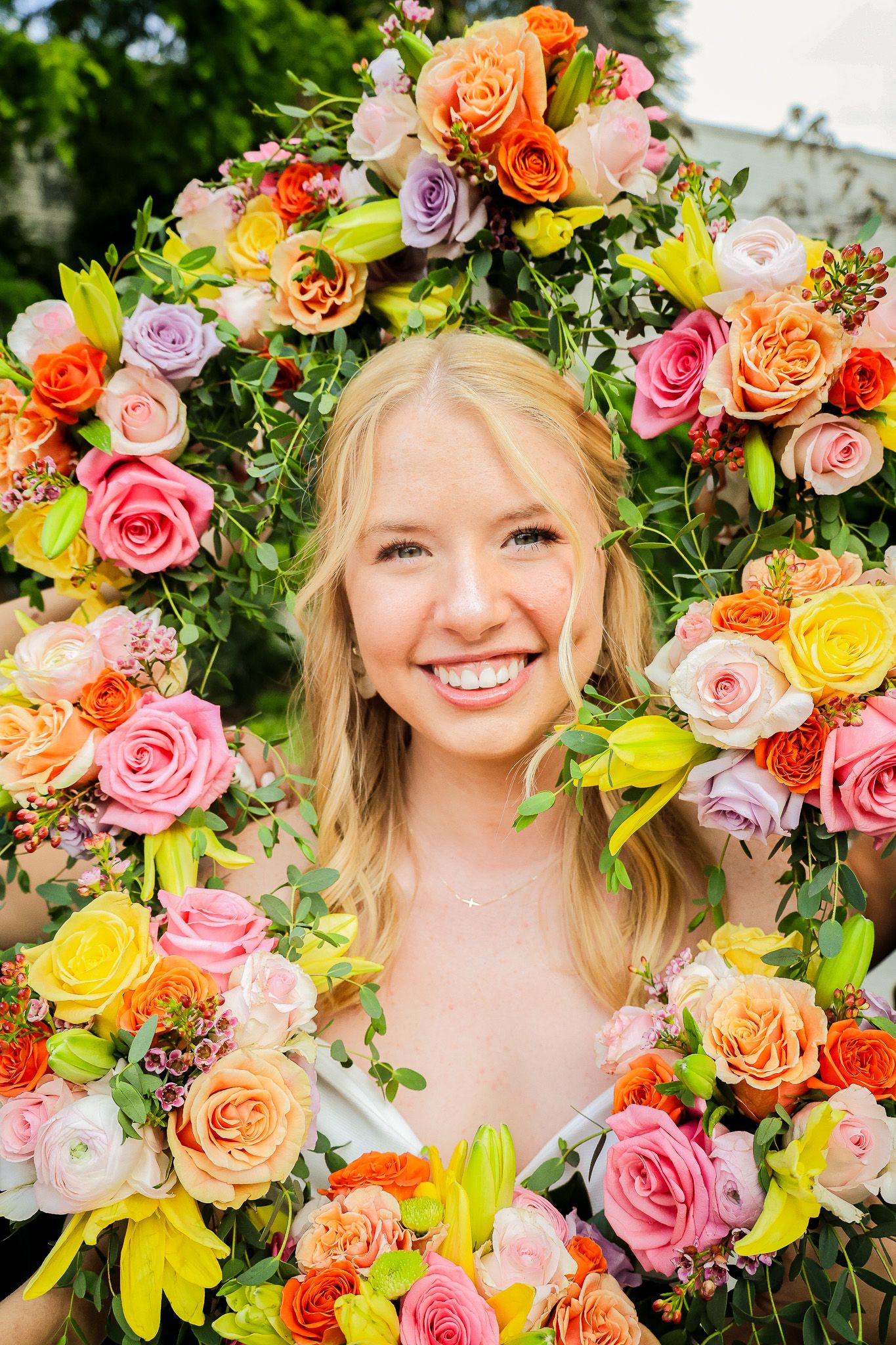 Smiling blonde bride framed by vibrant colorful floral wreath of roses and mixed blooms outdoors.