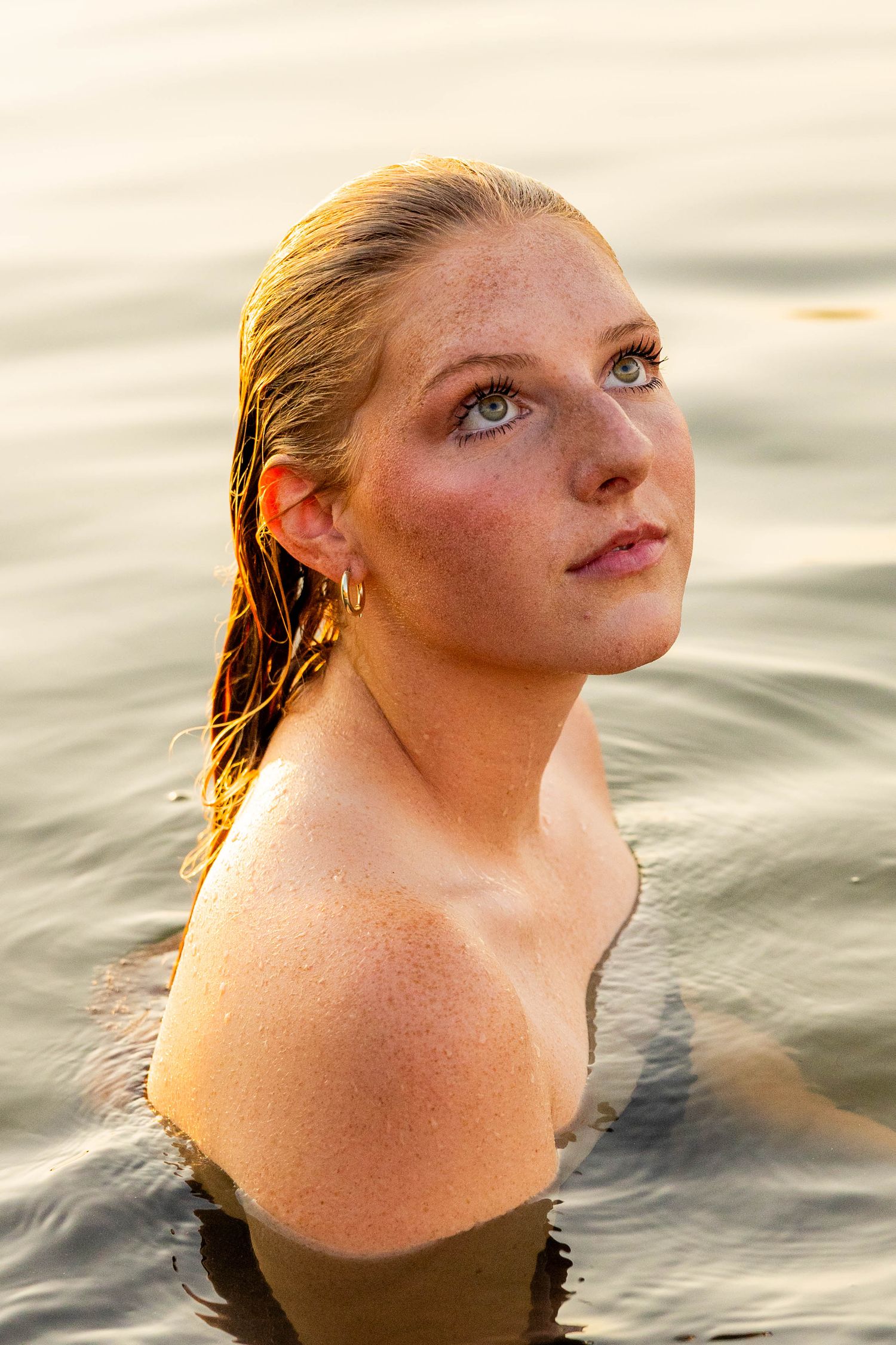Young woman with wet hair and freckles emerging from calm water, bathed in warm golden sunset light.