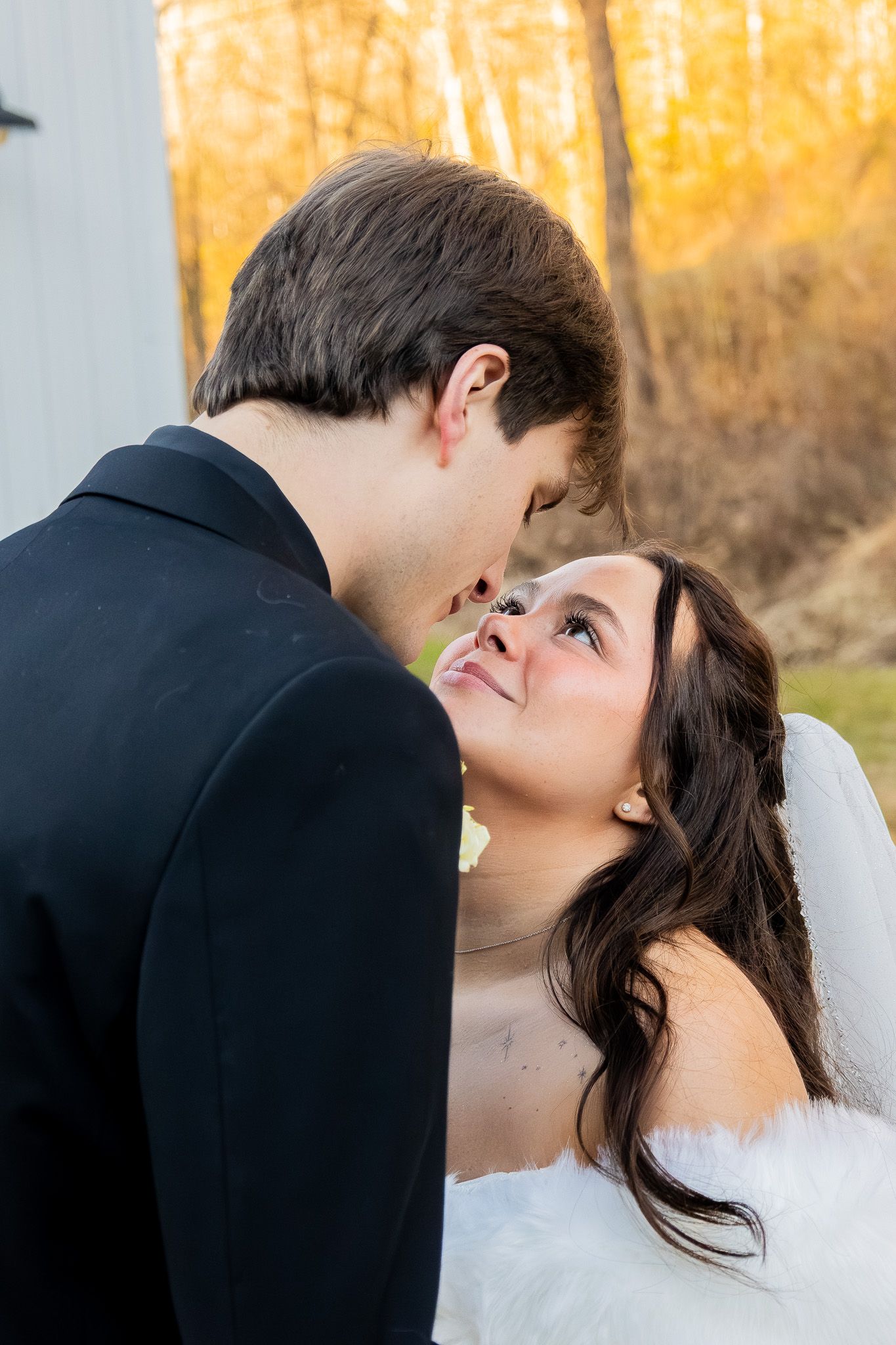 Bride and groom sharing a romantic moment, woman in white dress gazing up at man in black suit with autumn trees behind.