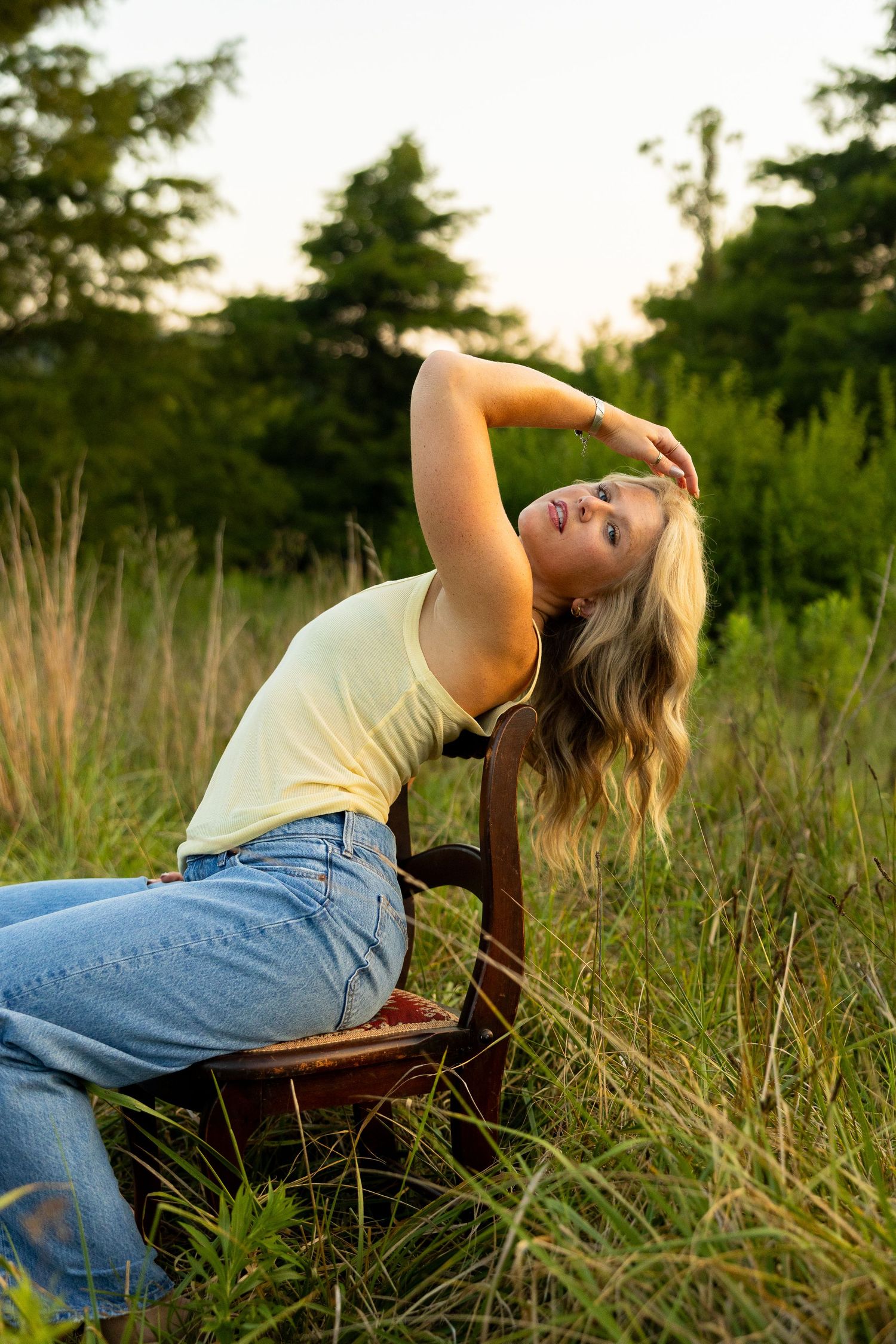Young blonde woman in yellow top and jeans poses gracefully on vintage chair in golden hour meadow.