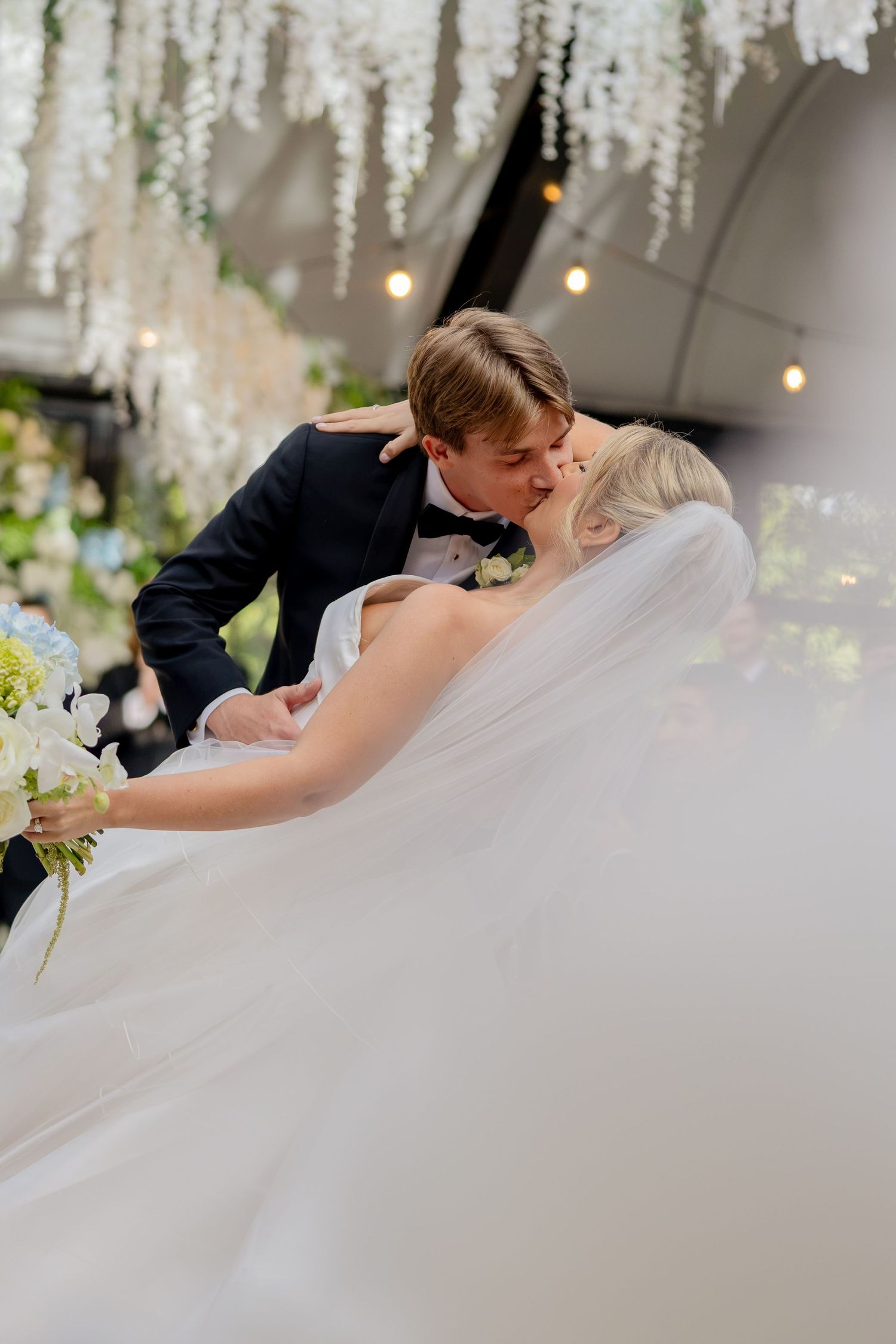 Bride and groom share a romantic dip kiss at their wedding ceremony, surrounded by white floral decorations and string lights.