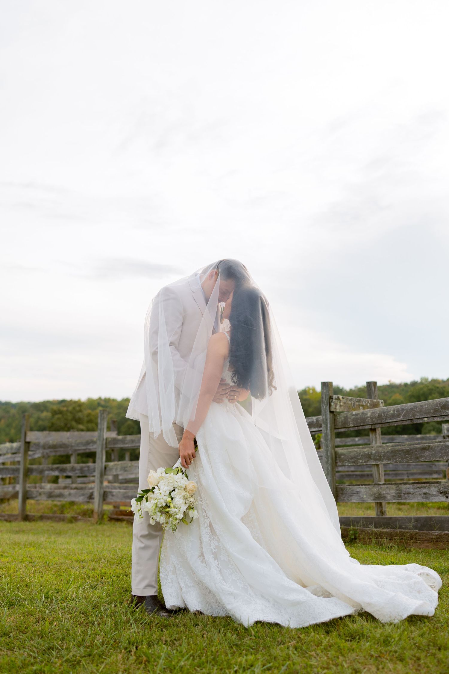 Bride and groom kissing under wedding veil outdoors near rustic wooden fence on green lawn.