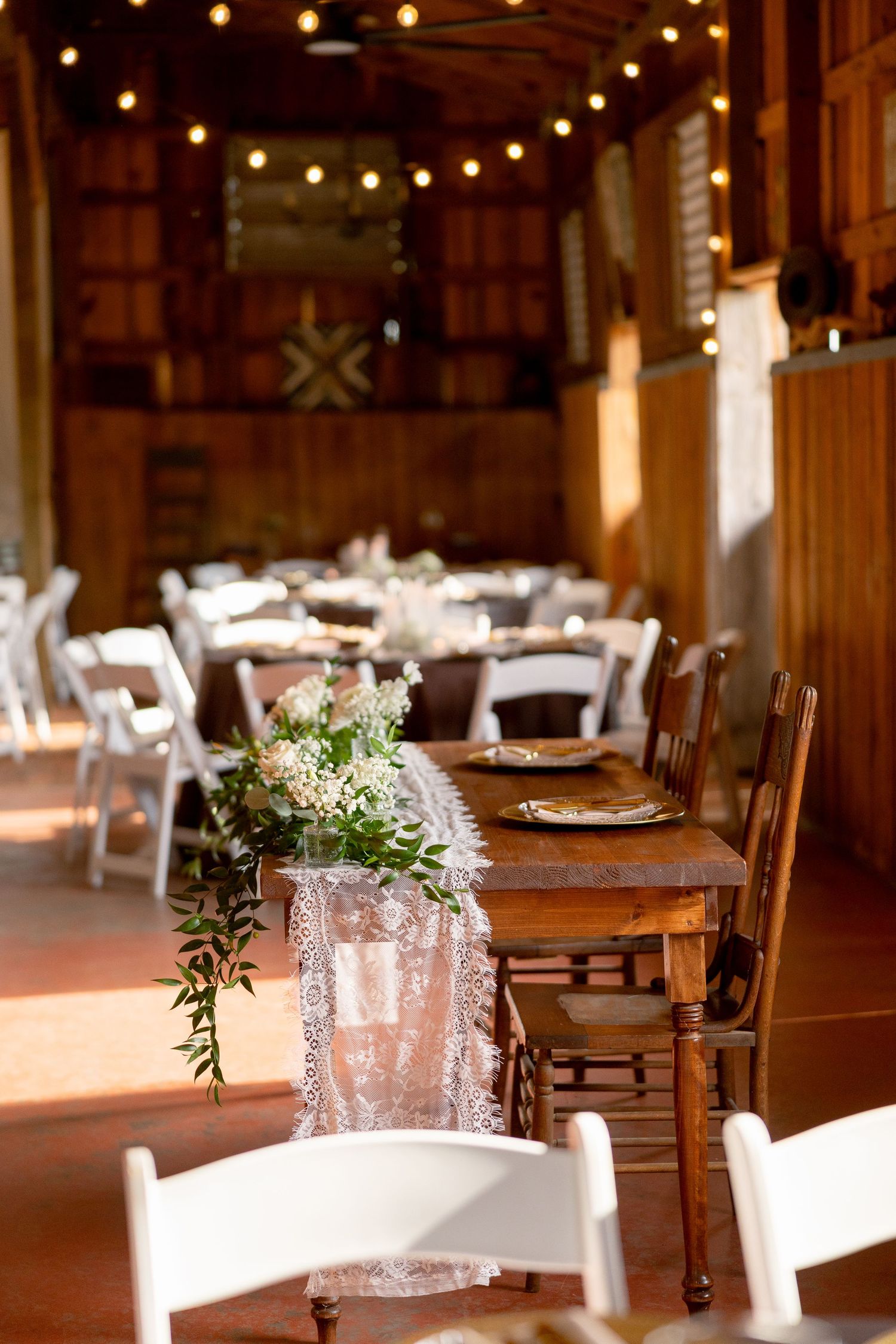 Rustic barn wedding reception with lace table runner, floral centerpieces, white folding chairs, and warm string lights overhead.