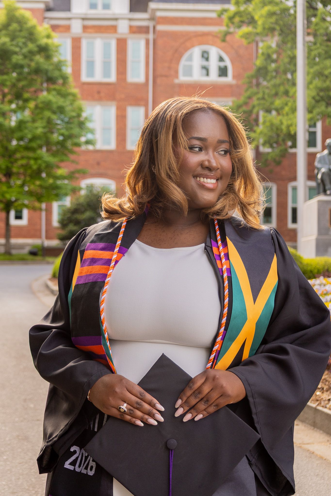 Smiling Black woman in graduation gown and kente stole holds cap outside brick university building, class of 2023.