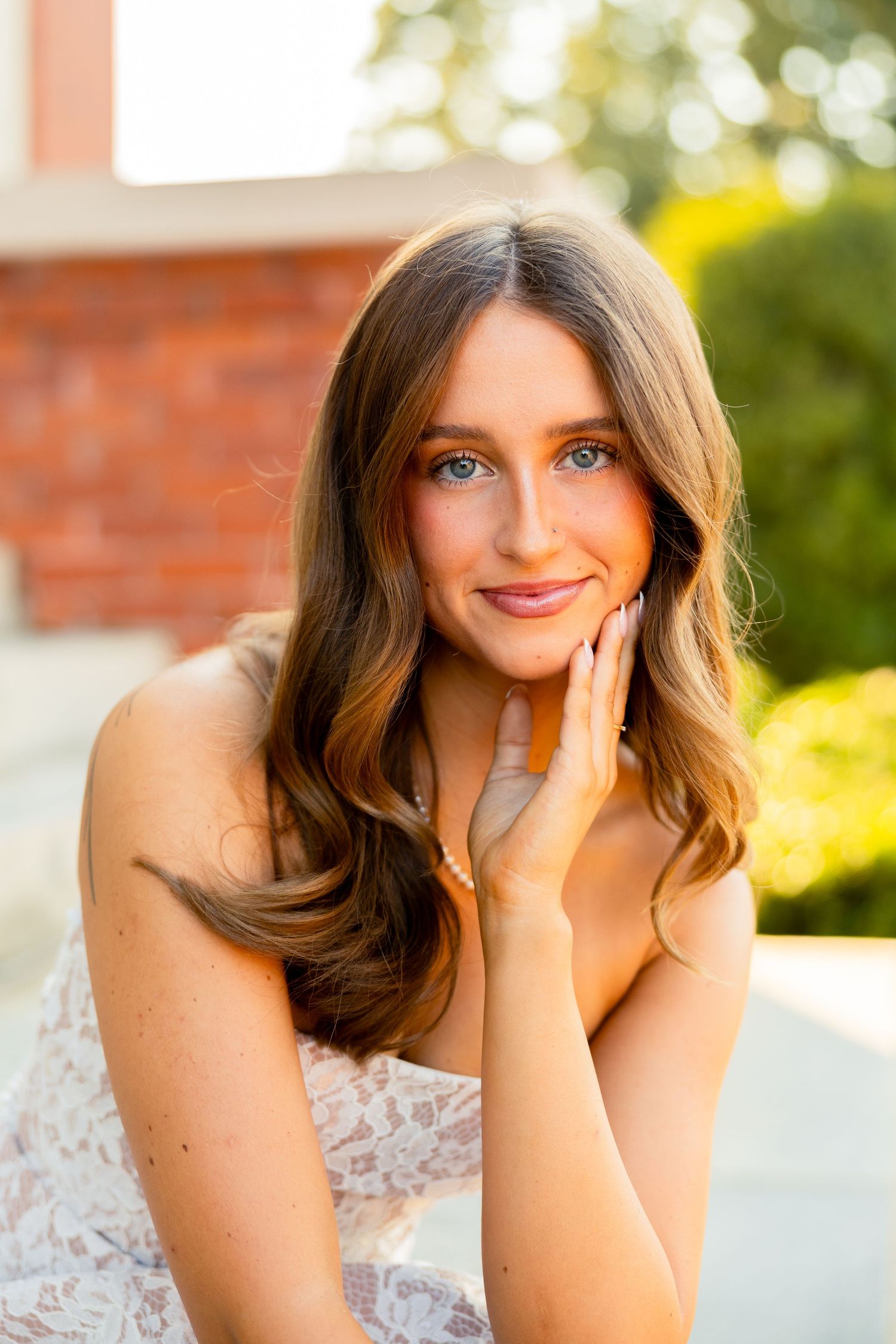 Young woman with long brown hair and blue eyes smiling softly, wearing a lace dress outdoors in warm sunlight.